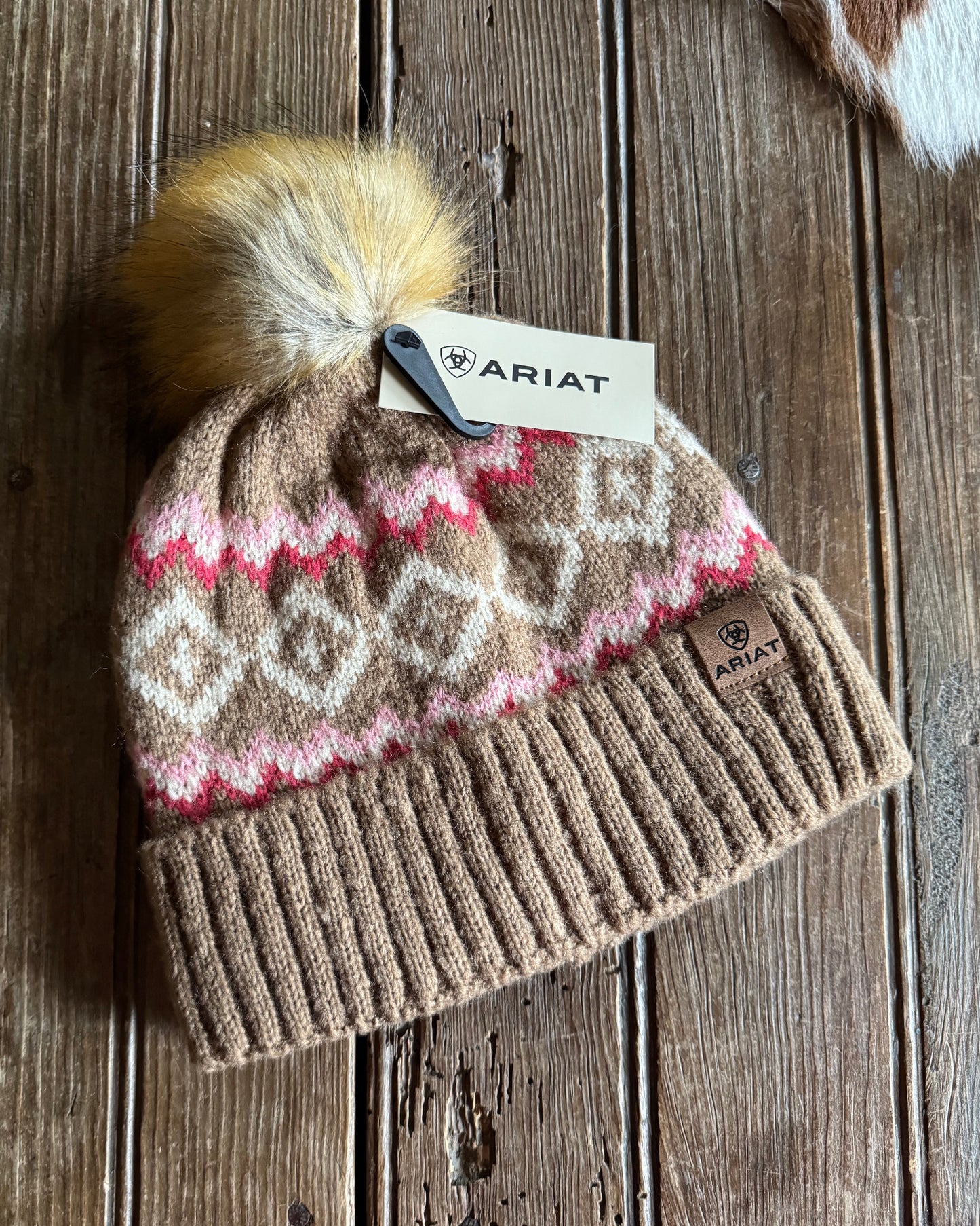 Knitted beanie with patterned front and pom-pom on a wooden surface, Ariat brand tag visible