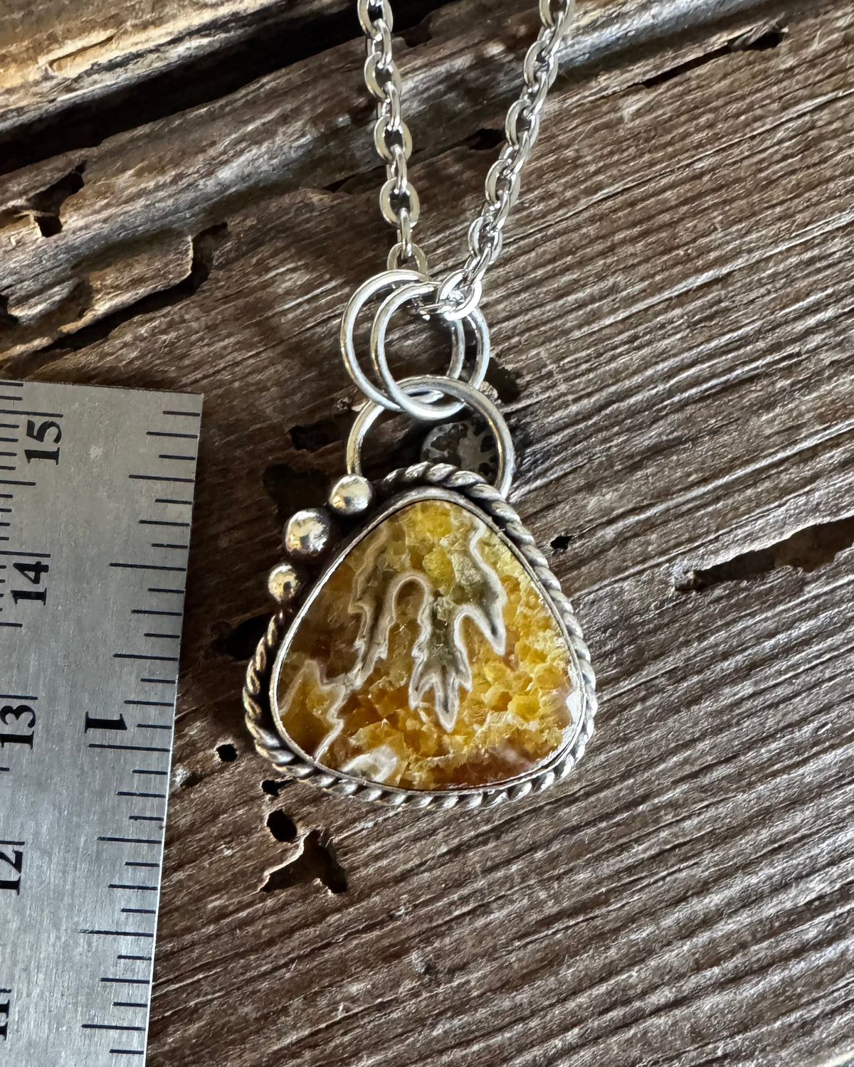 A silver pendant necklace with a triangular agate stone pendant, displayed next to a stainless steel ruler on a rustic wooden surface.