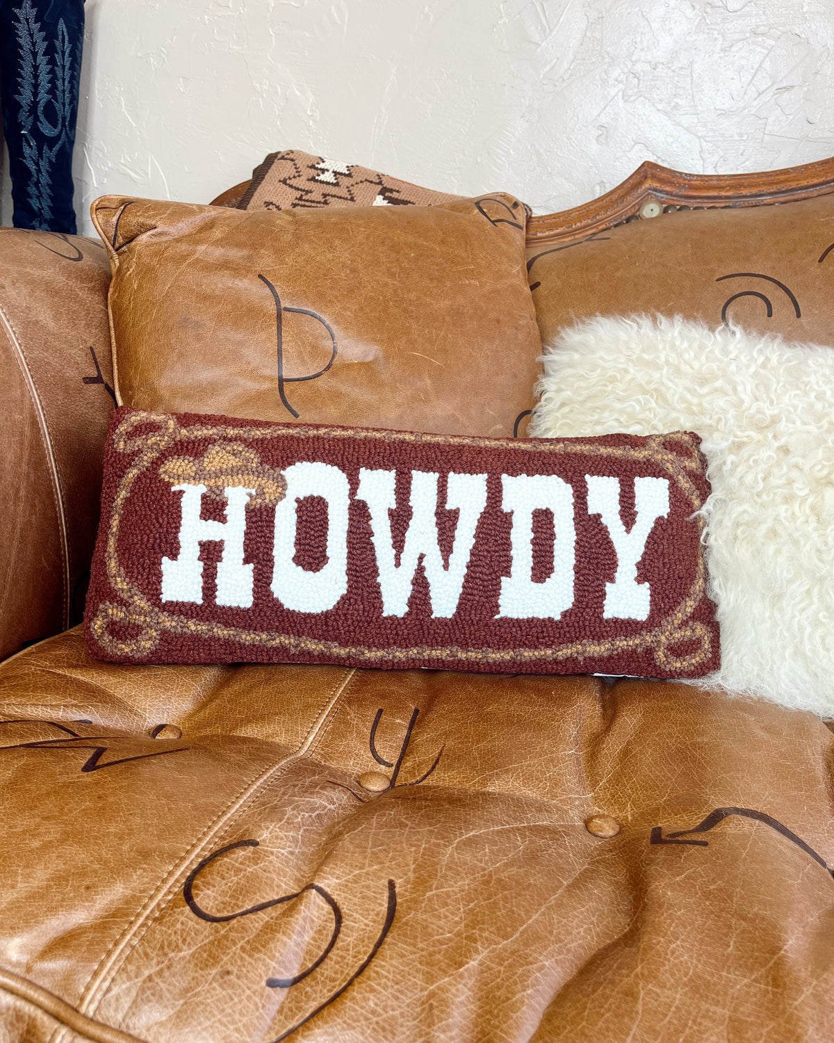 A rectangular woven throw pillow with the word 'HOWDY' in white varsity letters on a dark red background, styled on a brown leather sofa next to a white faux fur pillow.