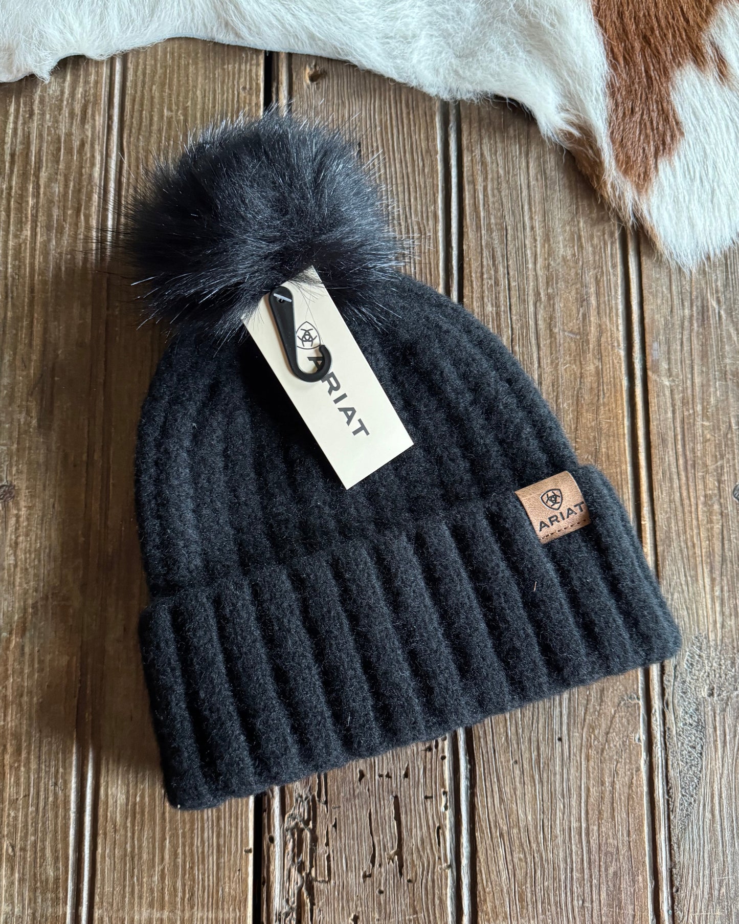 Black knit beanie with pom-pom on a wooden surface, featuring a brand tag.
