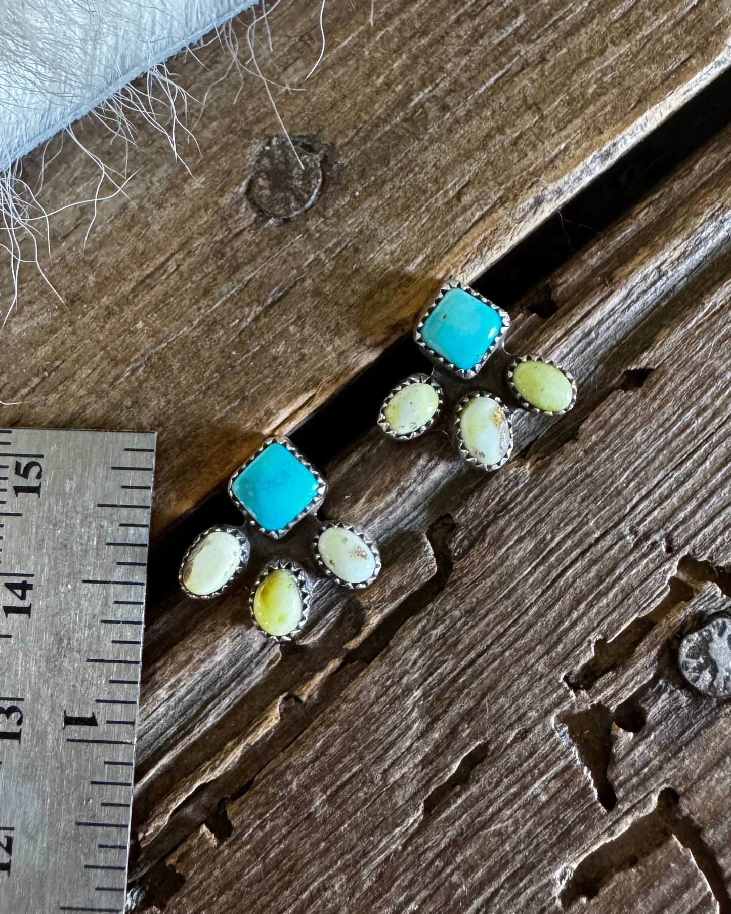 A close-up of several small jewelry pieces laid on a weathered wooden surface, with a metal ruler and white fur visible for scale. The jewelry appears to include turquoise-blue and pale yellow stones in silver-toned settings.