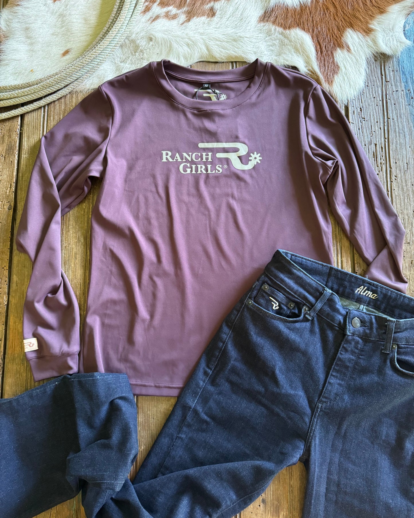 Purple long-sleeve shirt with 'Ranch Girls' logo and blue denim jacket on a wooden surface.
