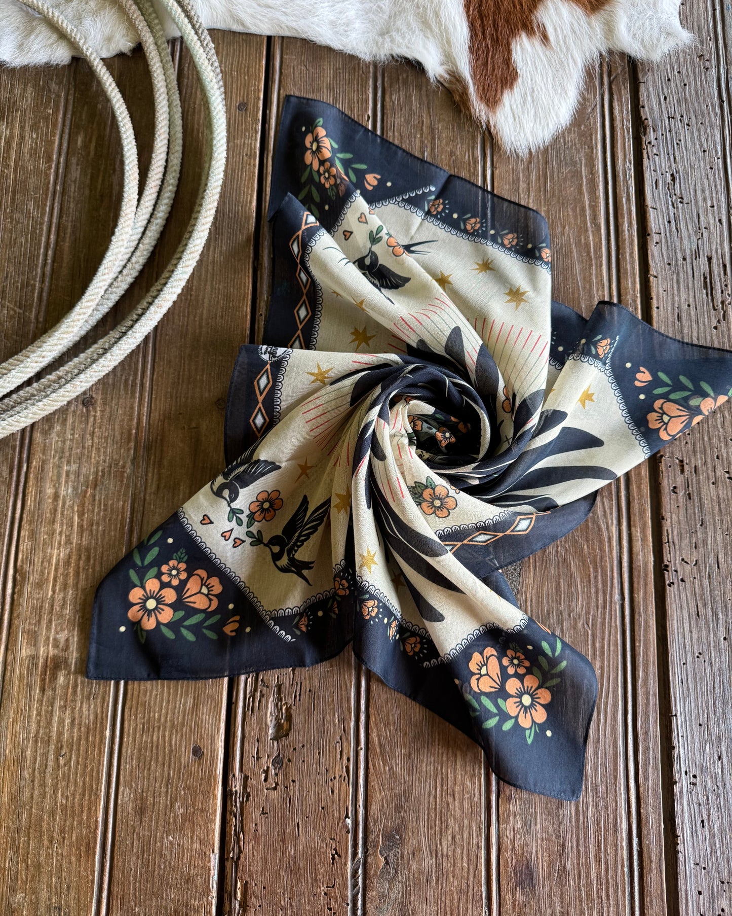 Decorative scarf with floral patterns on a wooden surface next to a cowhide rug and rope.