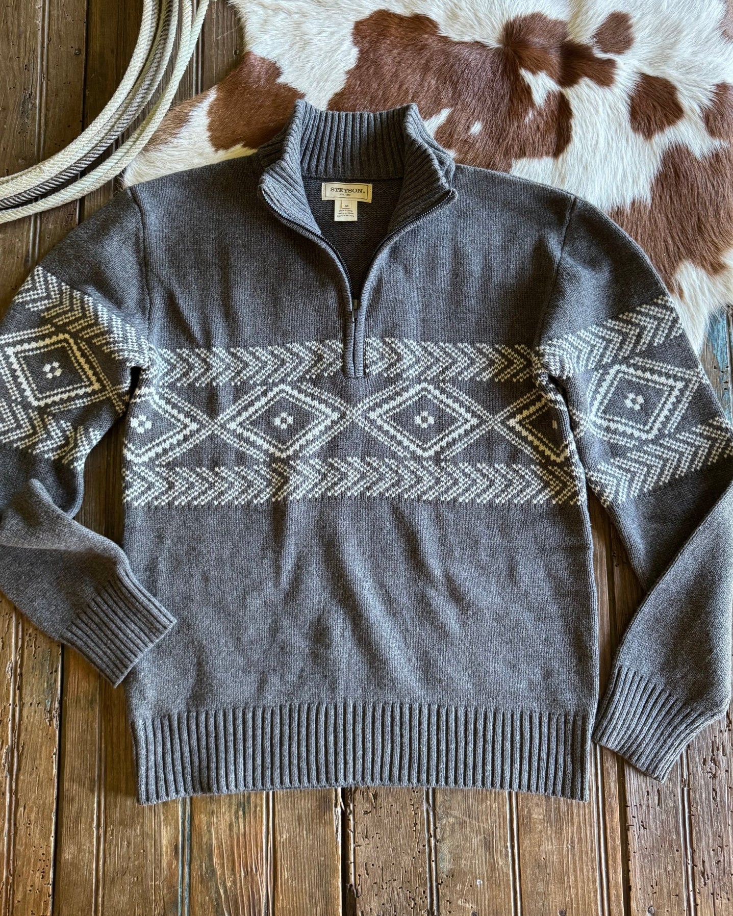 Gray sweater with geometric pattern on a wooden surface next to a cowhide rug and lasso.