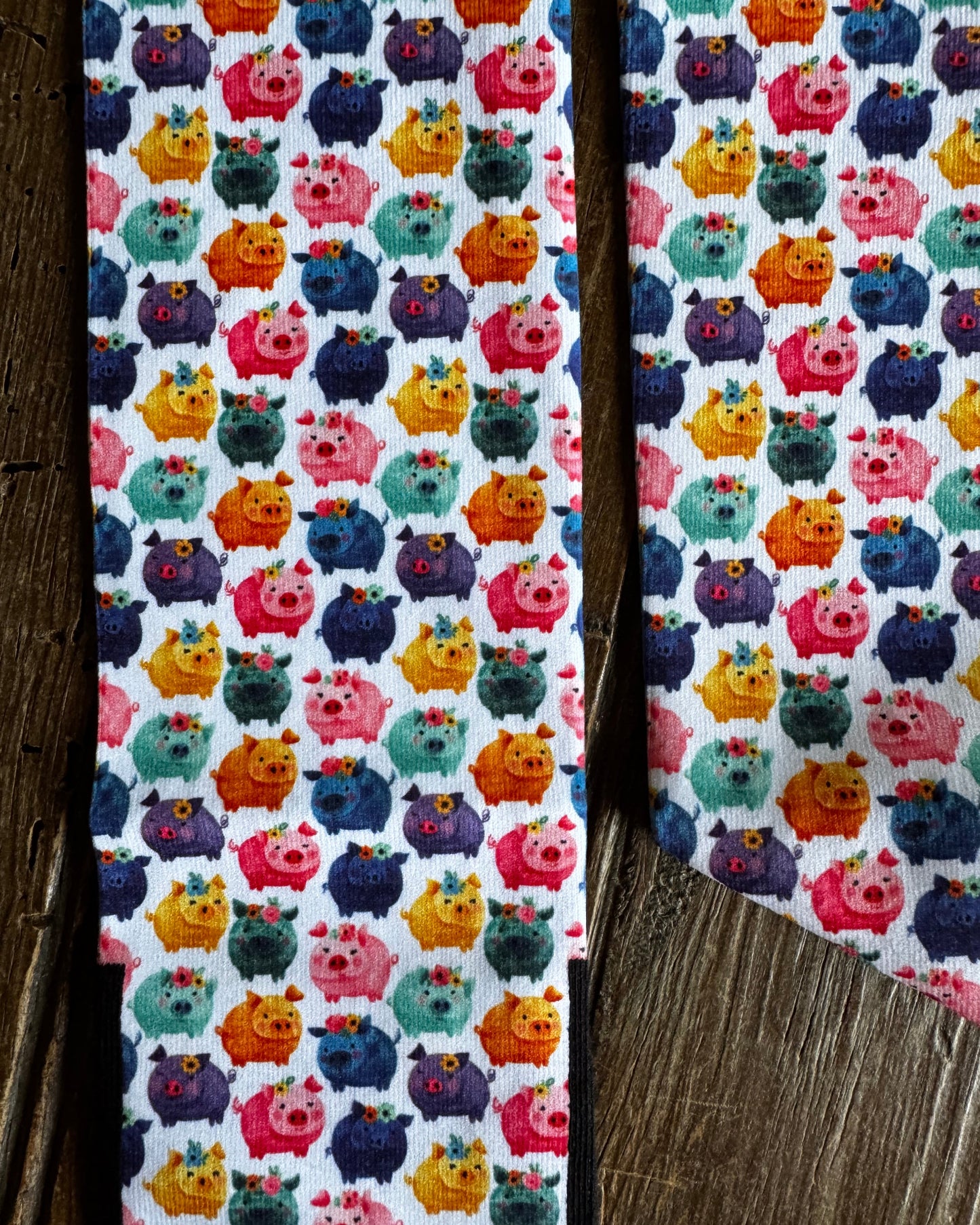 A close-up of a white fabric with a colorful repeating pattern of cartoon pig-like creatures in pink, orange, yellow, teal, and purple, laid flat on a rustic wooden surface.