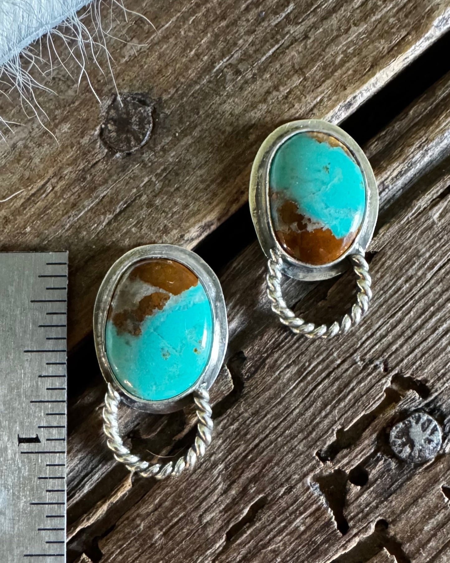 A pair of silver-toned drop earrings with oval turquoise stones and twisted rope details, displayed on a rustic wooden surface next to a metal ruler for scale.