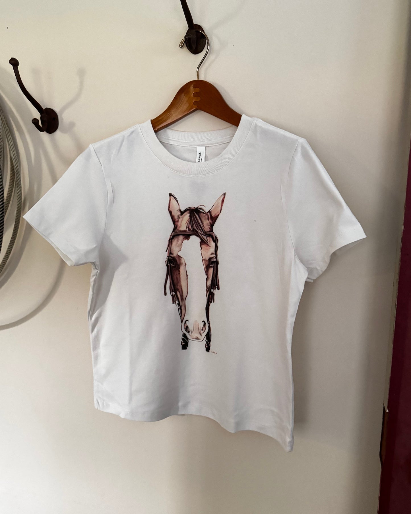 A white short-sleeve t-shirt hanging on a wooden hanger against a beige wall, featuring a graphic print of a horse head wearing a lasso.