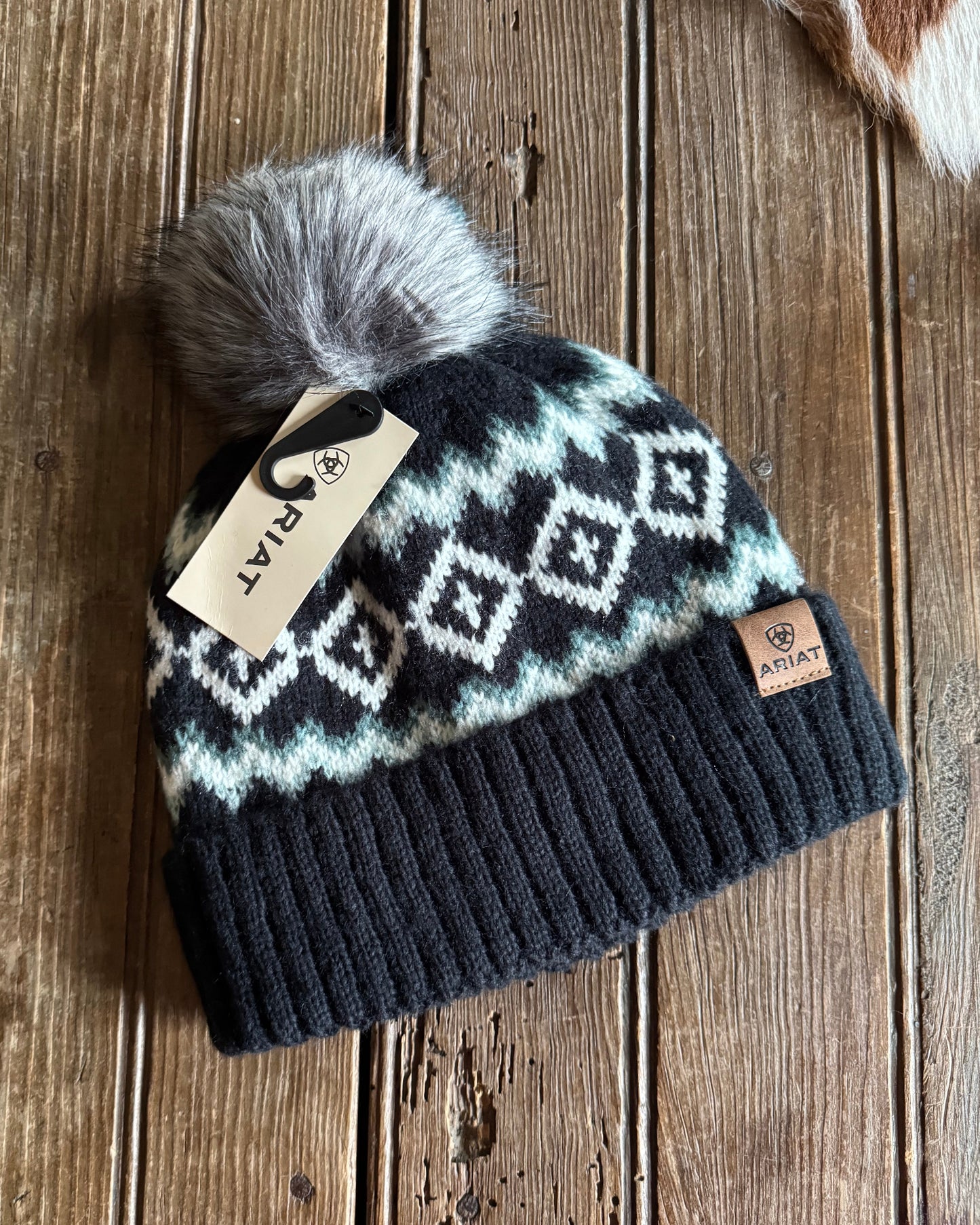 Knitted beanie with pattern and pom-pom on a wooden surface, featuring a brand tag.