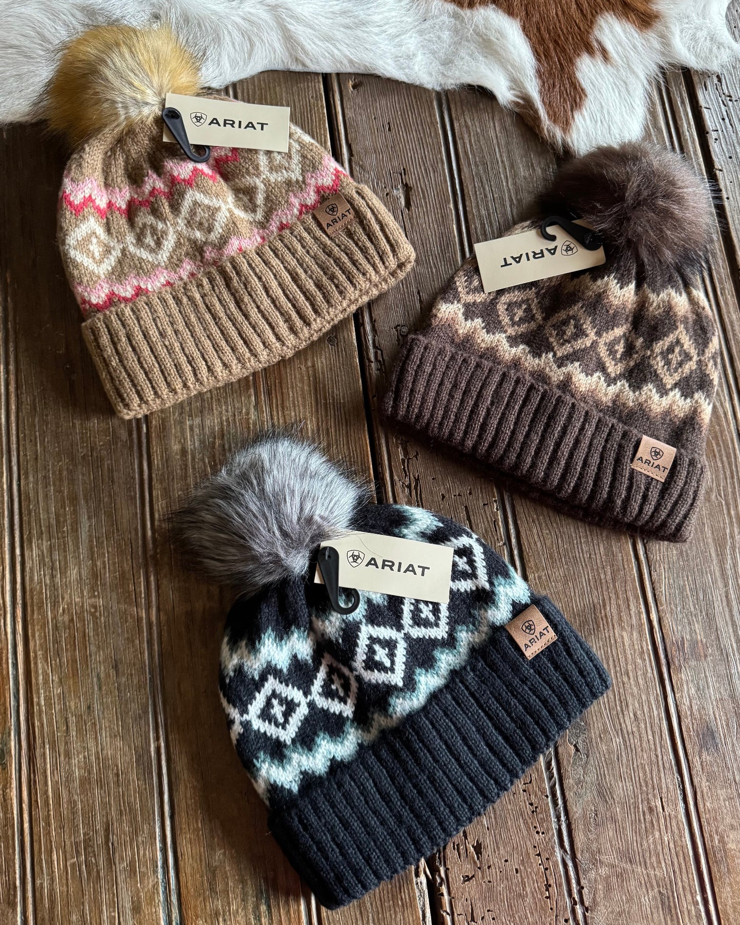 Three knit hats with patterned designs and pom-poms on a wooden surface, Ariat branded.