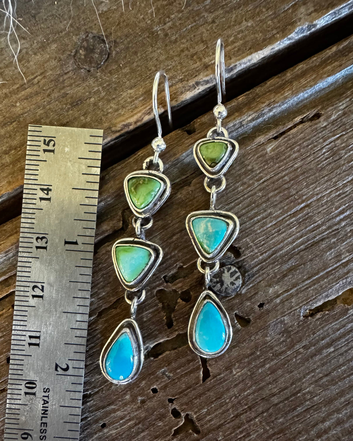A pair of silver drop earrings with green and turquoise stones, shown next to a metal ruler on a rustic wooden surface.
