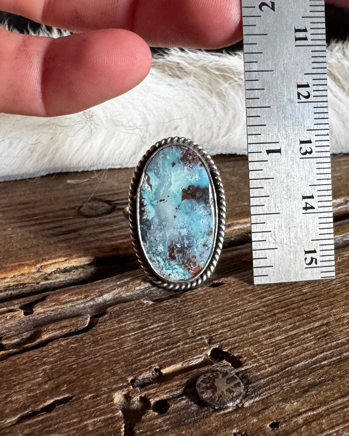 A hand holds a silver-toned ring with an oval turquoise-blue stone next to a stainless steel ruler for scale, set on a rustic wooden surface.