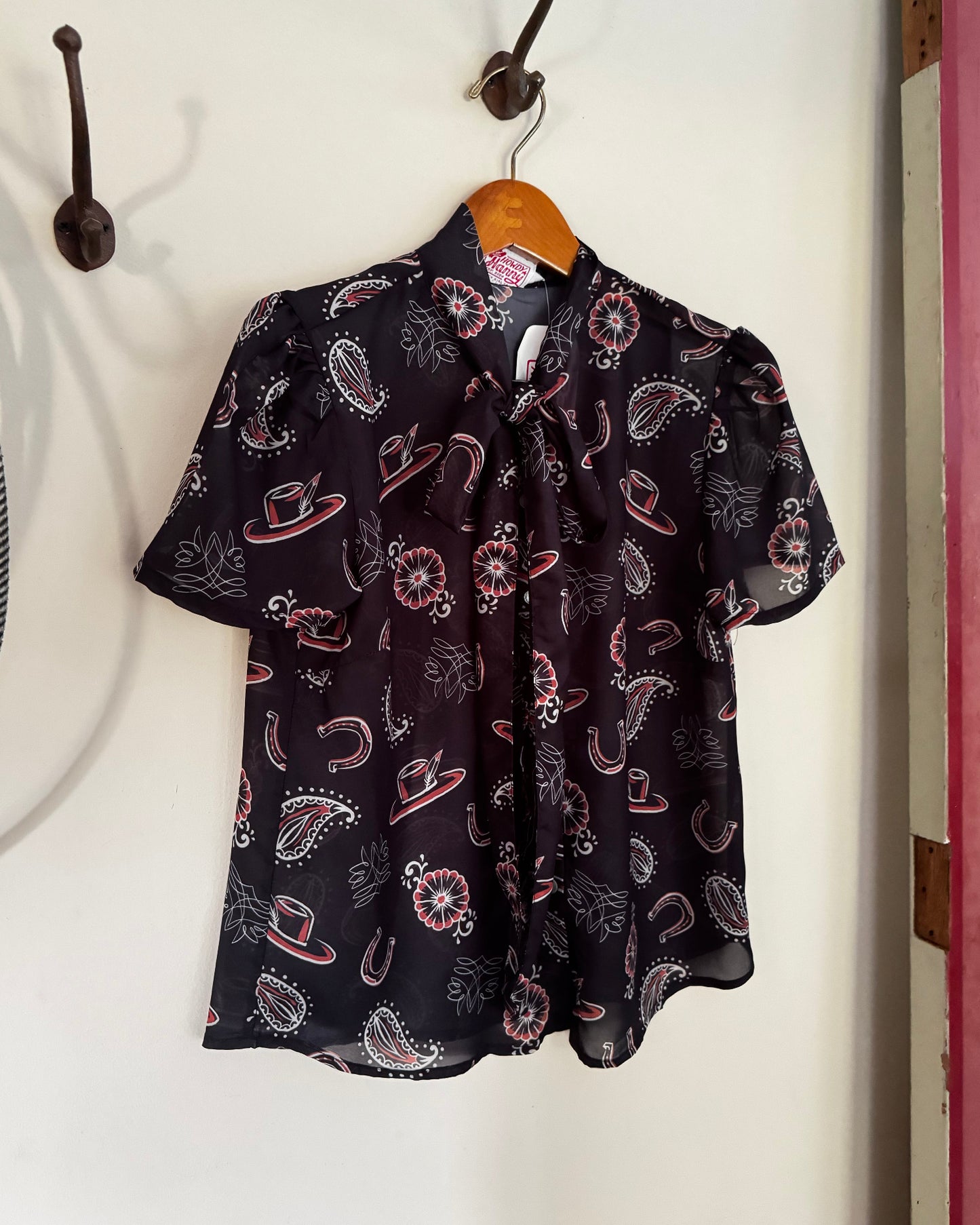 A short-sleeve black blouse with a paisley and hat pattern in pink, white, and red, hanging on a wooden hanger from a metal hook on a white wall.