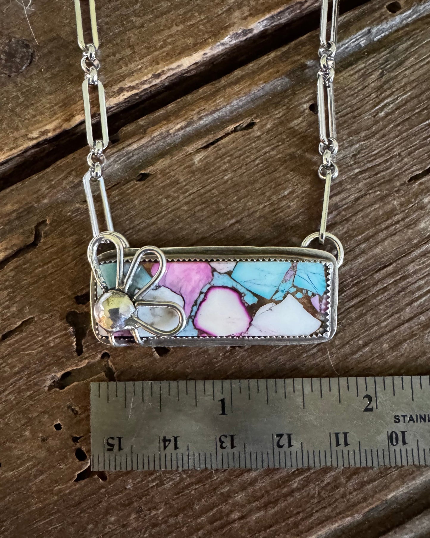 A silver-toned rectangular pendant necklace with a colorful marbled inlay of pink, blue, white, and brown, displayed on a rustic wooden surface next to a 2-inch ruler for scale.