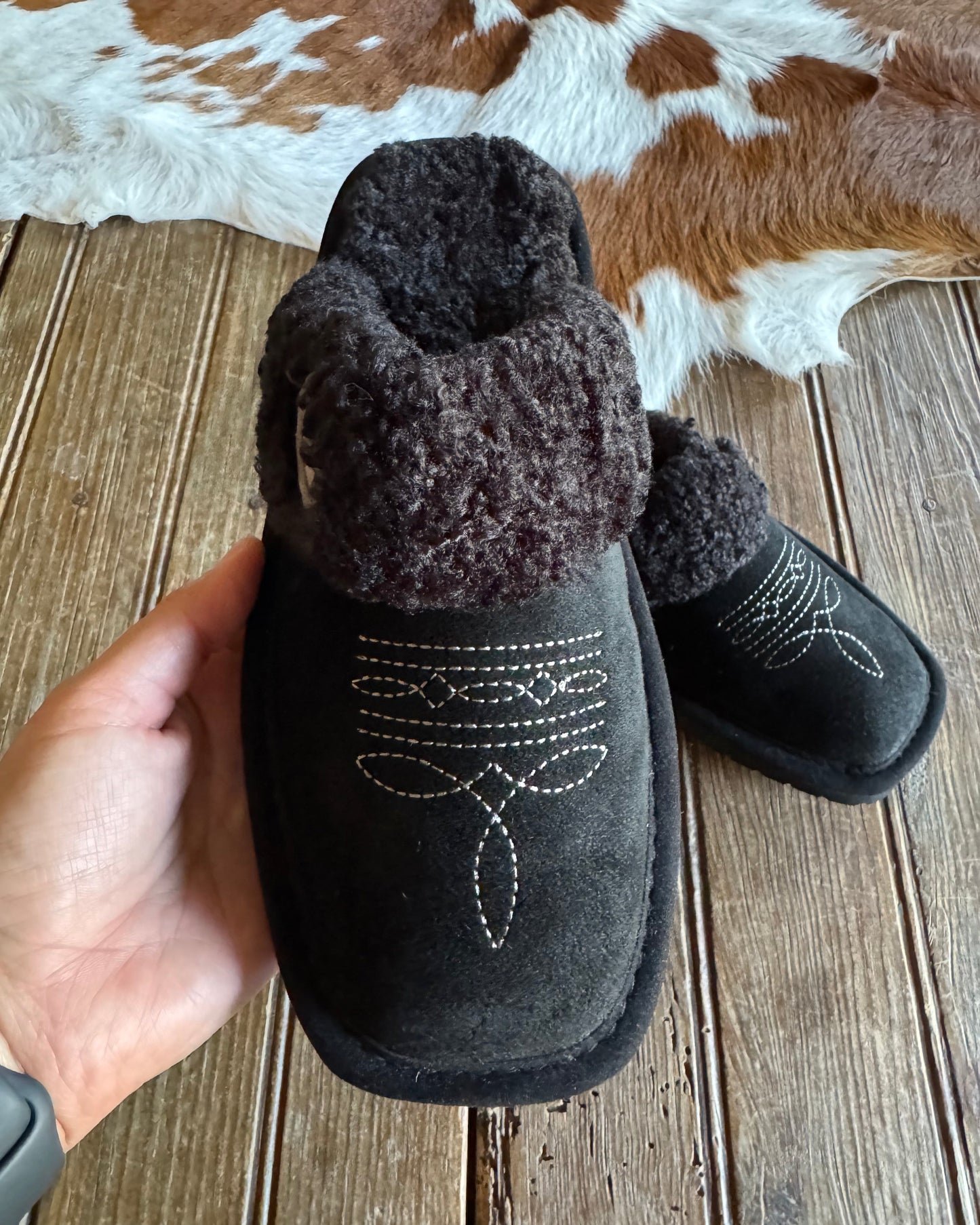 Black slippers with fuzzy trim held by a hand on a wooden floor.