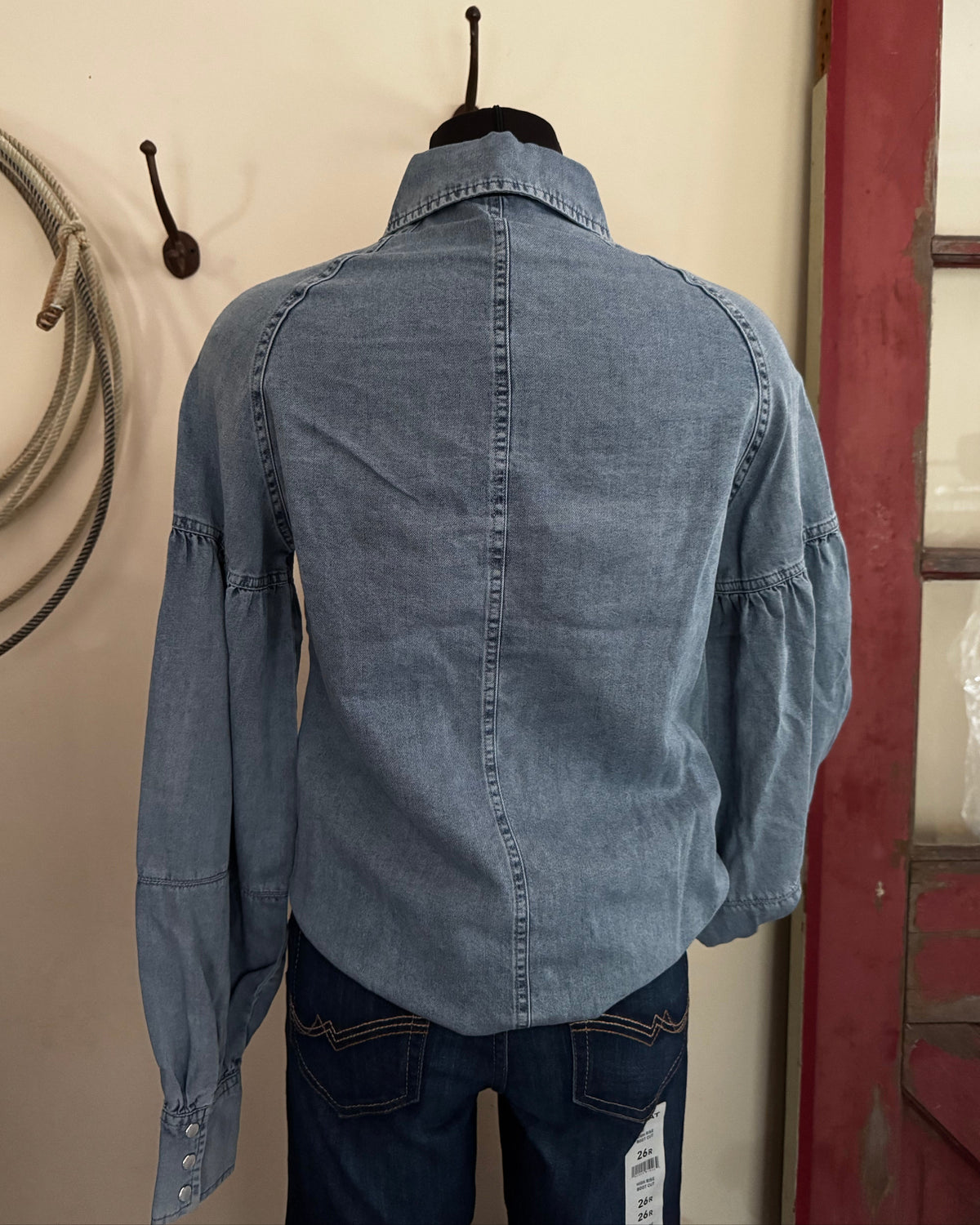 A light blue denim shirt-jacket with long puff sleeves, shown from the back on a mannequin torso with dark blue jeans. The jacket has a central seam down the back, gathered shoulder seams, and patch pockets.