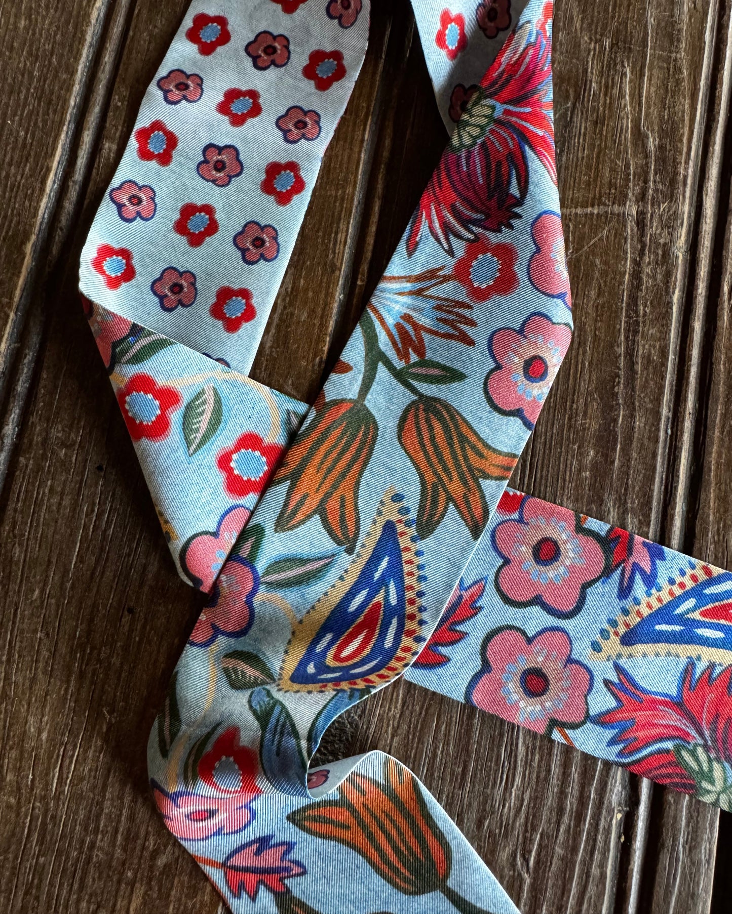 A light blue necktie with a dense floral and paisley print in red, pink, orange, and blue, laid flat on a dark wood background.