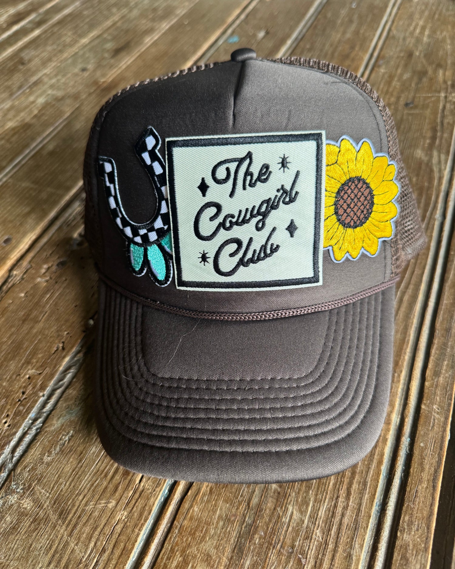 Brown foam trucker cap with sunflower design and 'The Cowgirl Club' text on a wooden surface