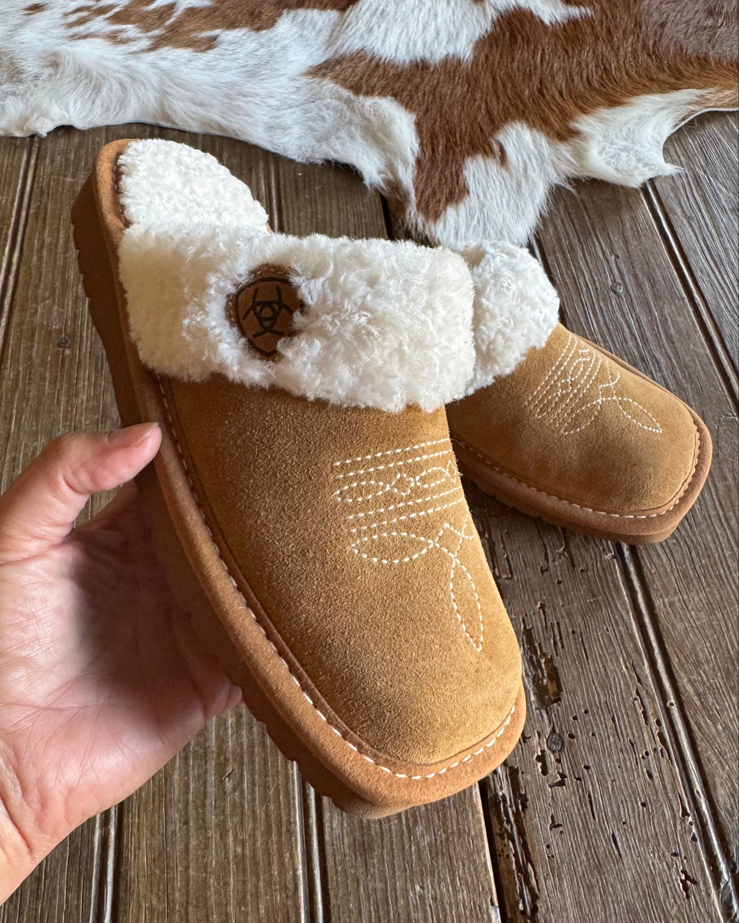 Brown slipper with white fur lining held by a hand on a wooden floor.