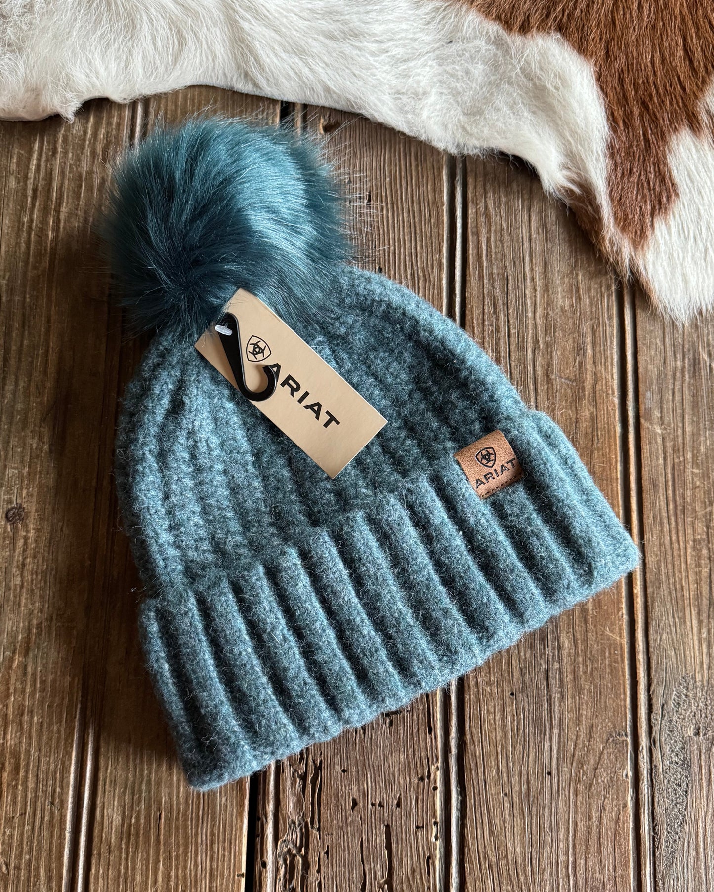 Blue knit beanie with pom-pom on a wooden surface, Ariat brand visible