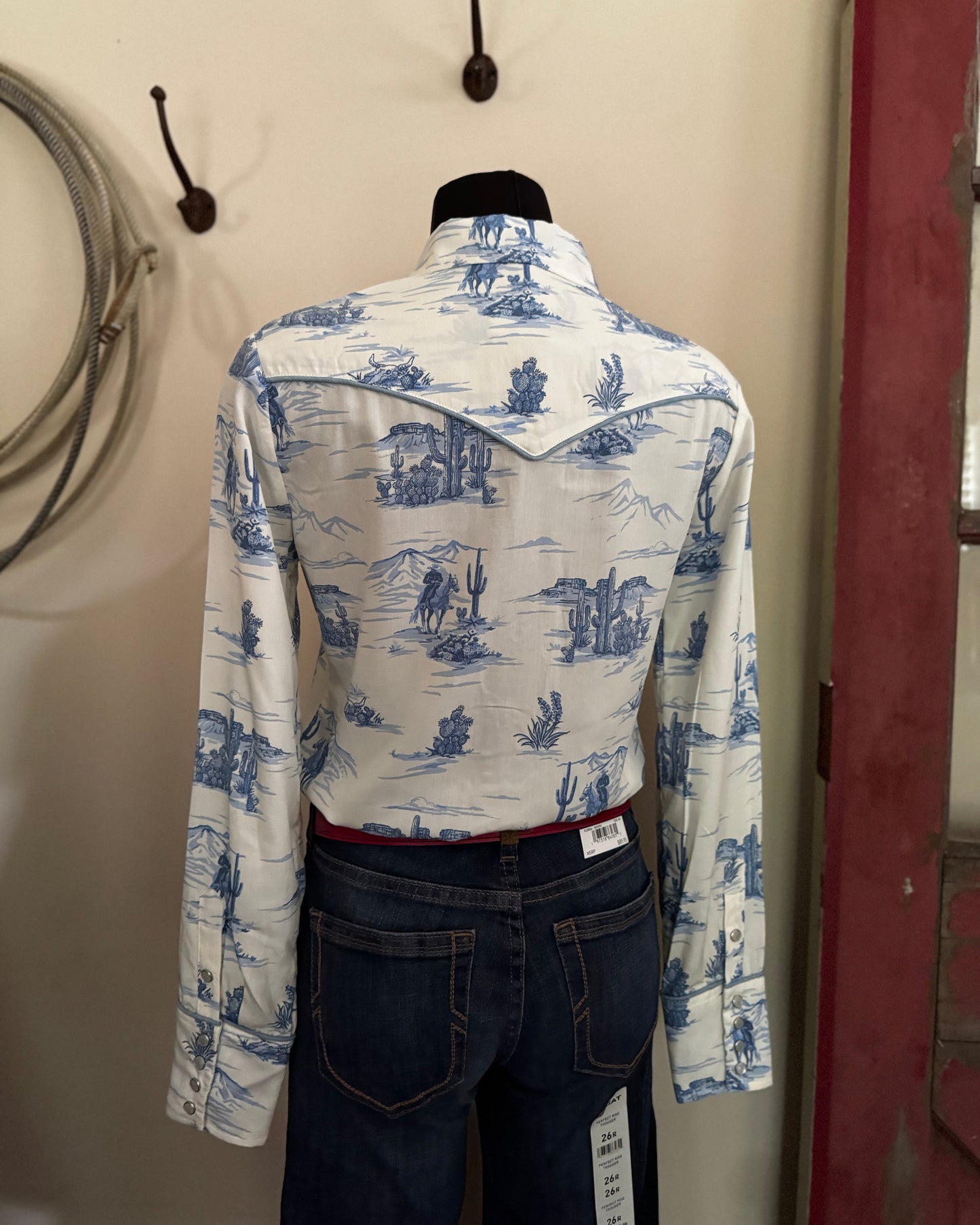 Mannequin wearing a white shirt with blue patterns and dark jeans against a neutral wall.