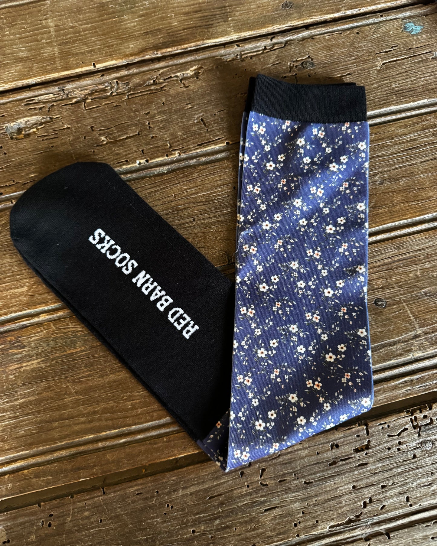 Black sock with 'Red Barn Socks' text and floral-patterned sock on a wooden surface