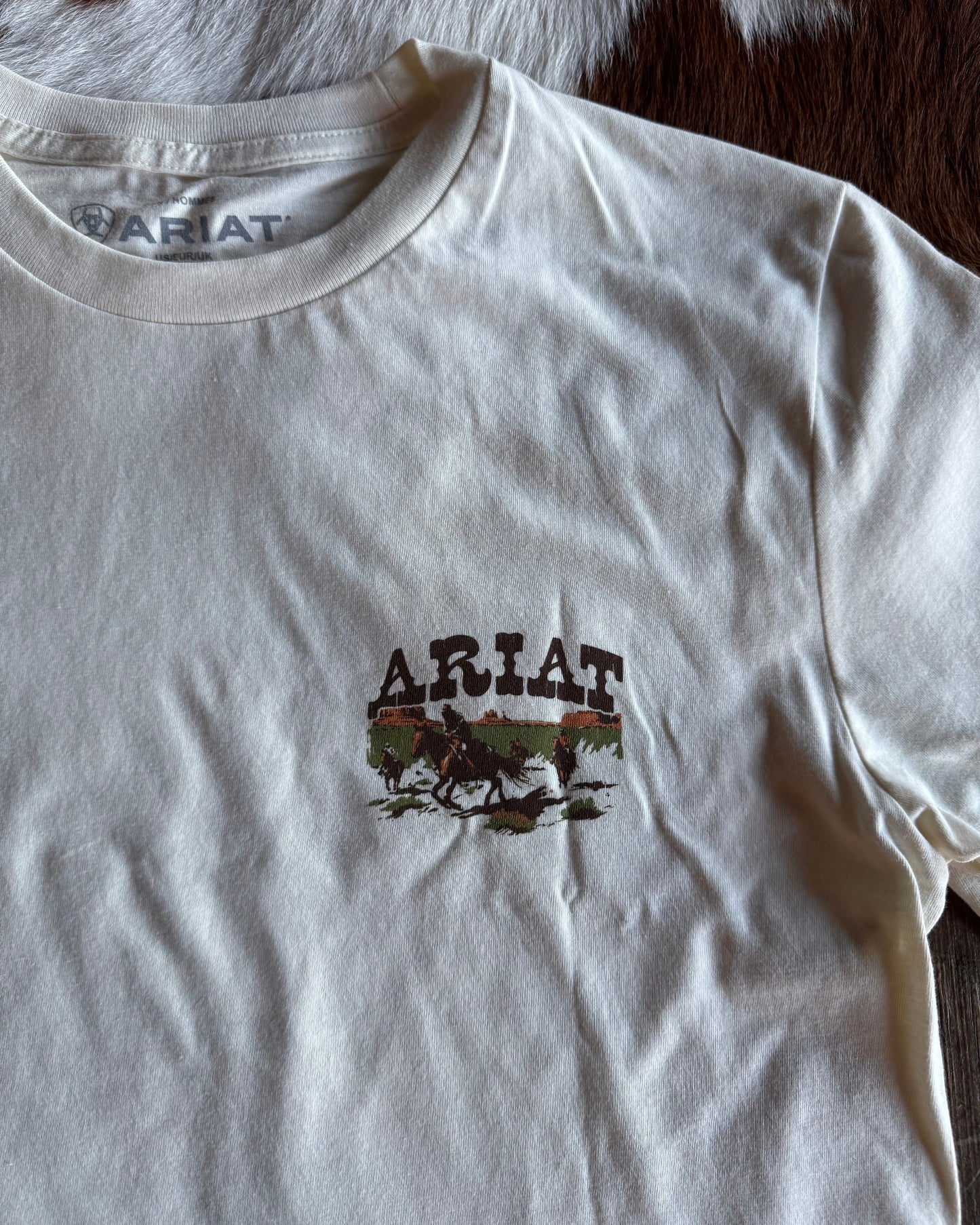 White t-shirt with Ariat logo and graphic on a brown and white cowhide rug