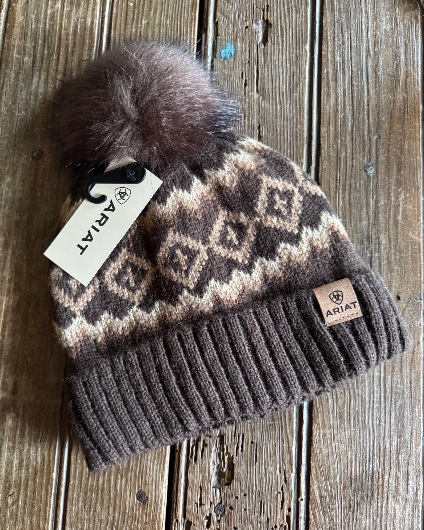 Brown beanie with patterned design and pom-pom on a wooden surface, featuring Ariat branding.