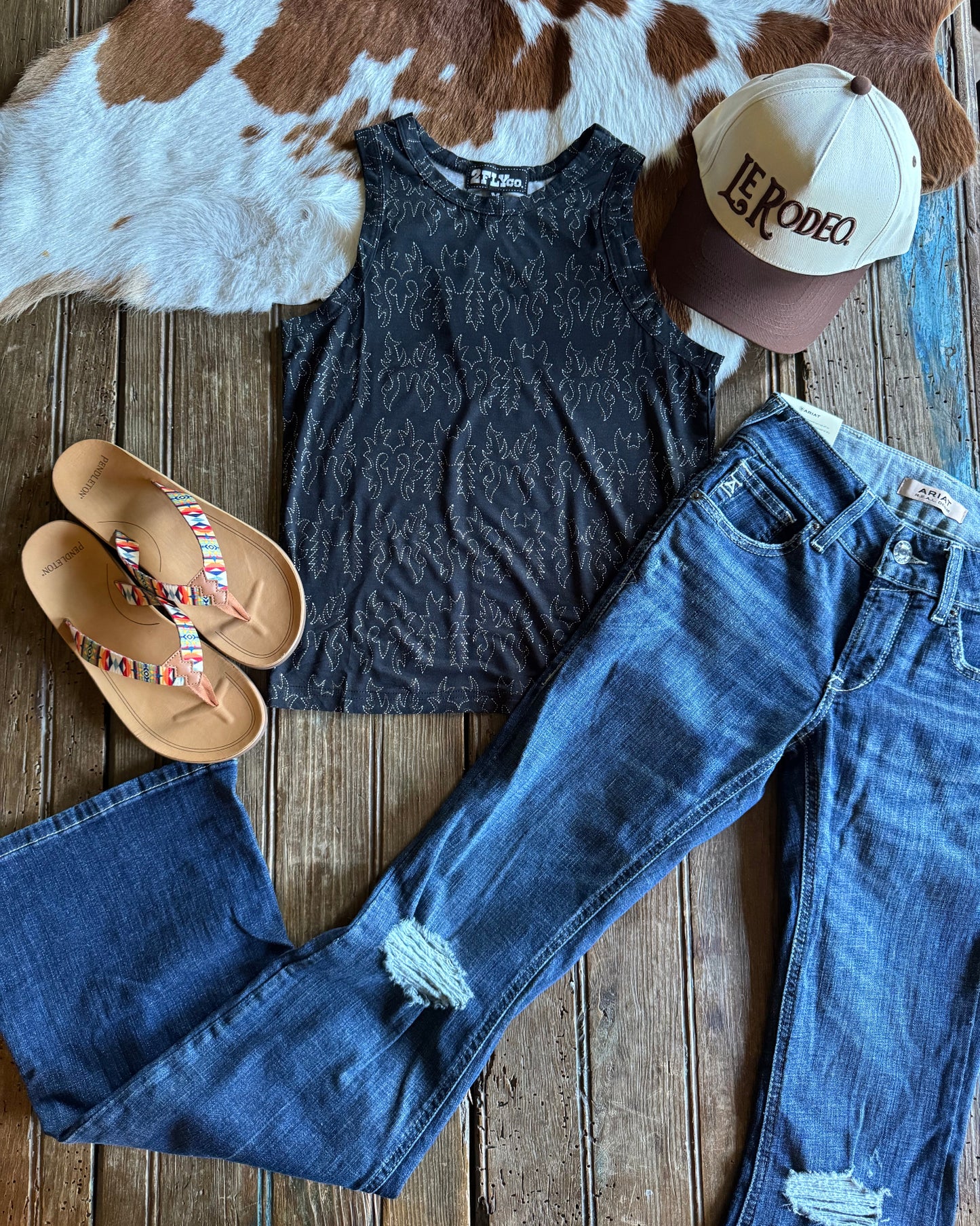 Blue jeans, black tank top, brown sandals, and a cap on a wooden surface with a cowhide rug.