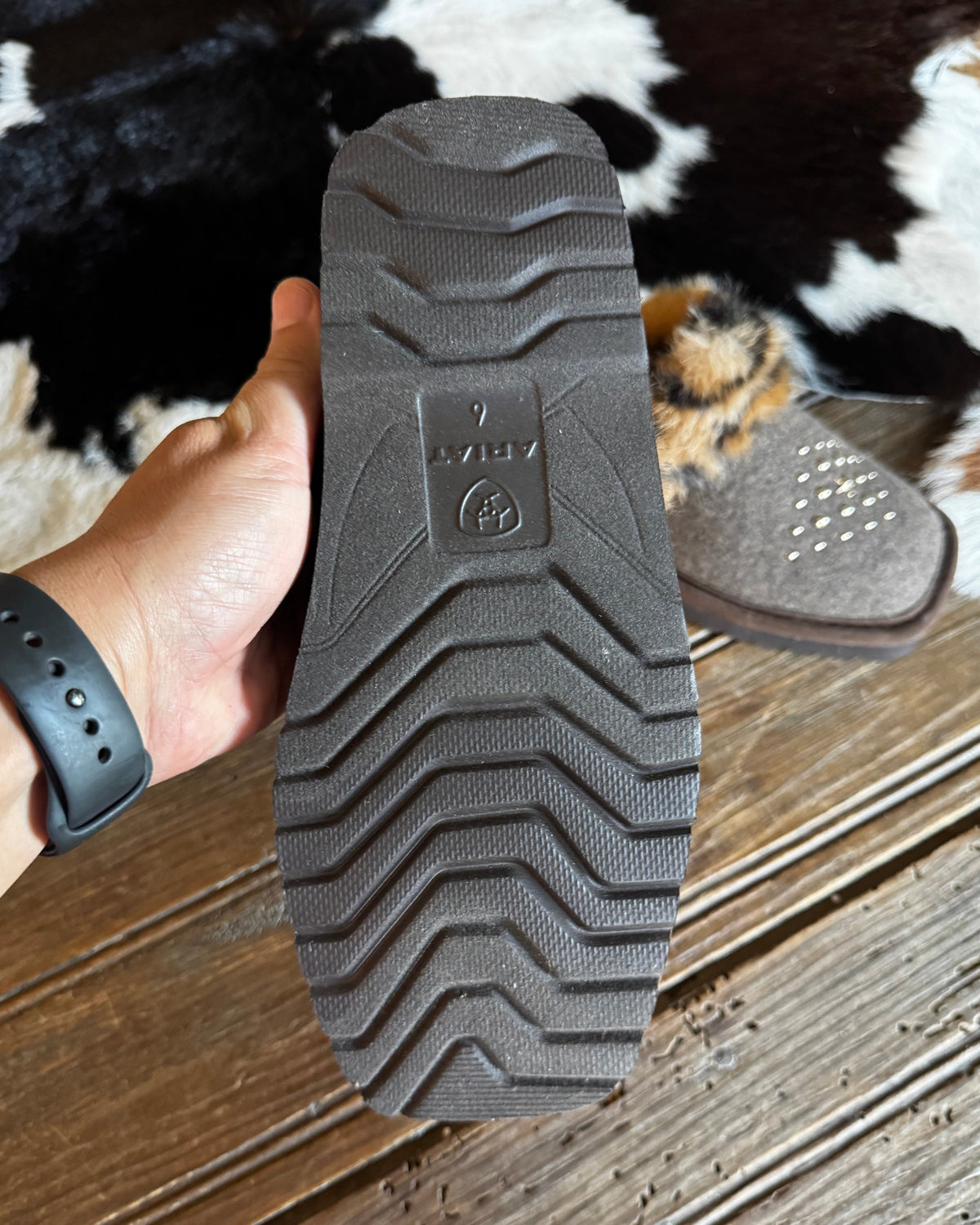 A hand holds up the treaded sole of a dark brown boot or shoe, showing the ARIAT brand logo embossed in the center. The background features a rustic wooden floor and a cowhide rug.