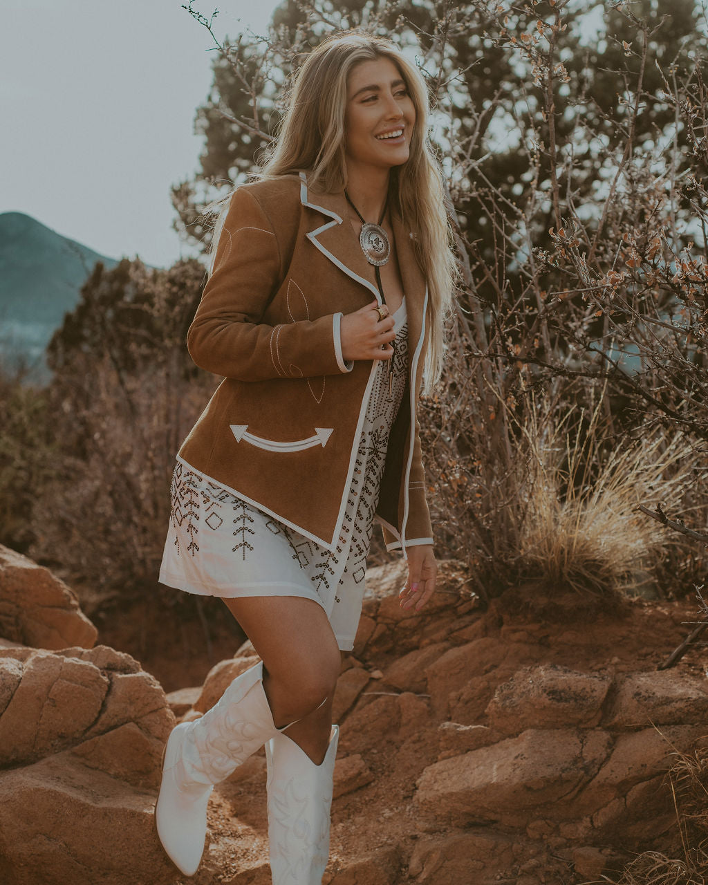 Woman in a brown jacket and white boots standing on rocky terrain with mountains in the background