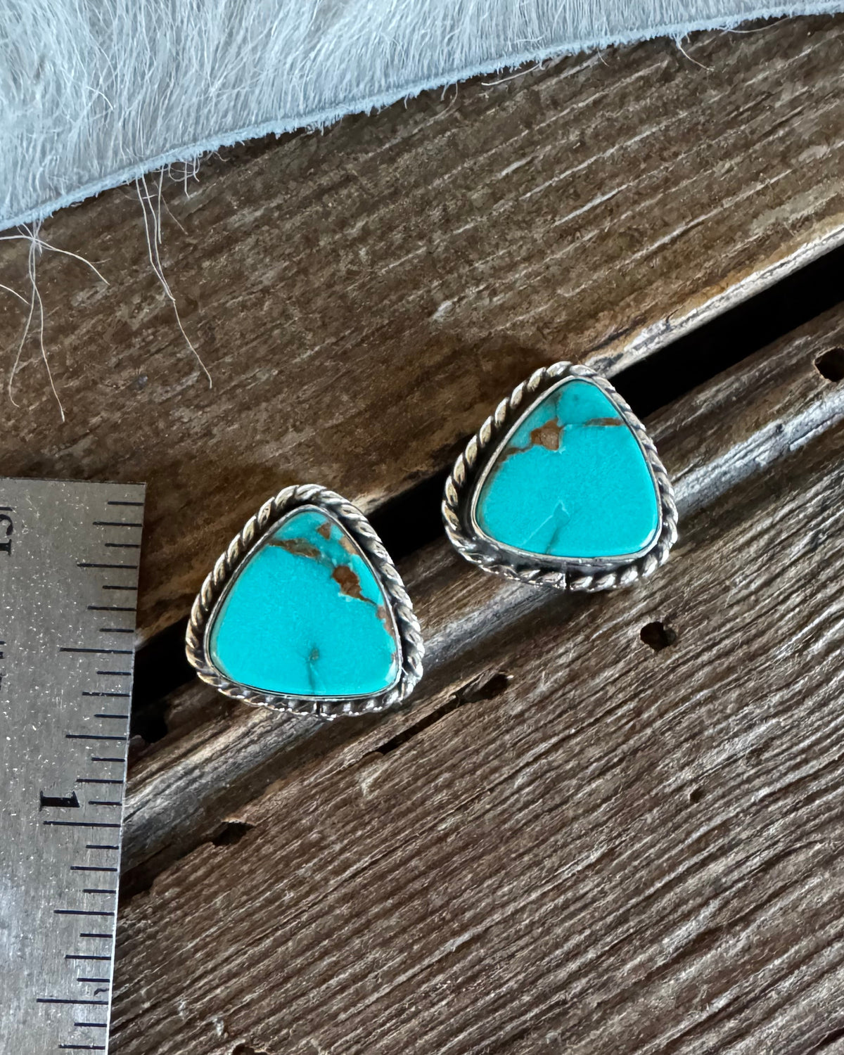A pair of Southwestern-style turquoise stud earrings with silver bezel settings and twisted rope borders, displayed on a rustic wooden surface next to a metal ruler for scale.