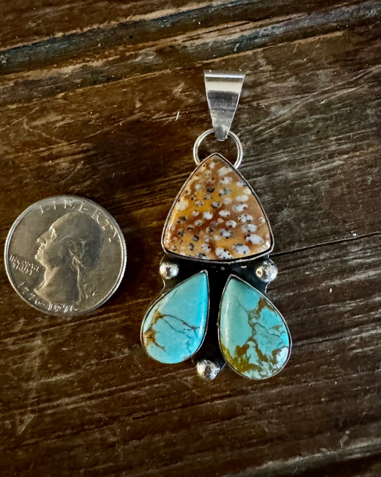 Turquoise and brown stone pendant with silver accents on a wooden surface, next to a quarter for scale.