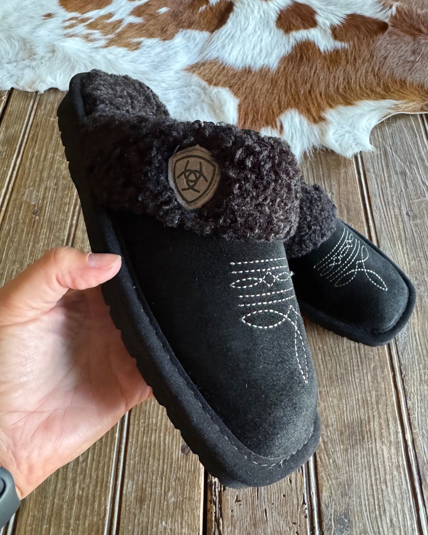 Black fuzzy slippers with a brand logo held by a hand on a wooden floor.