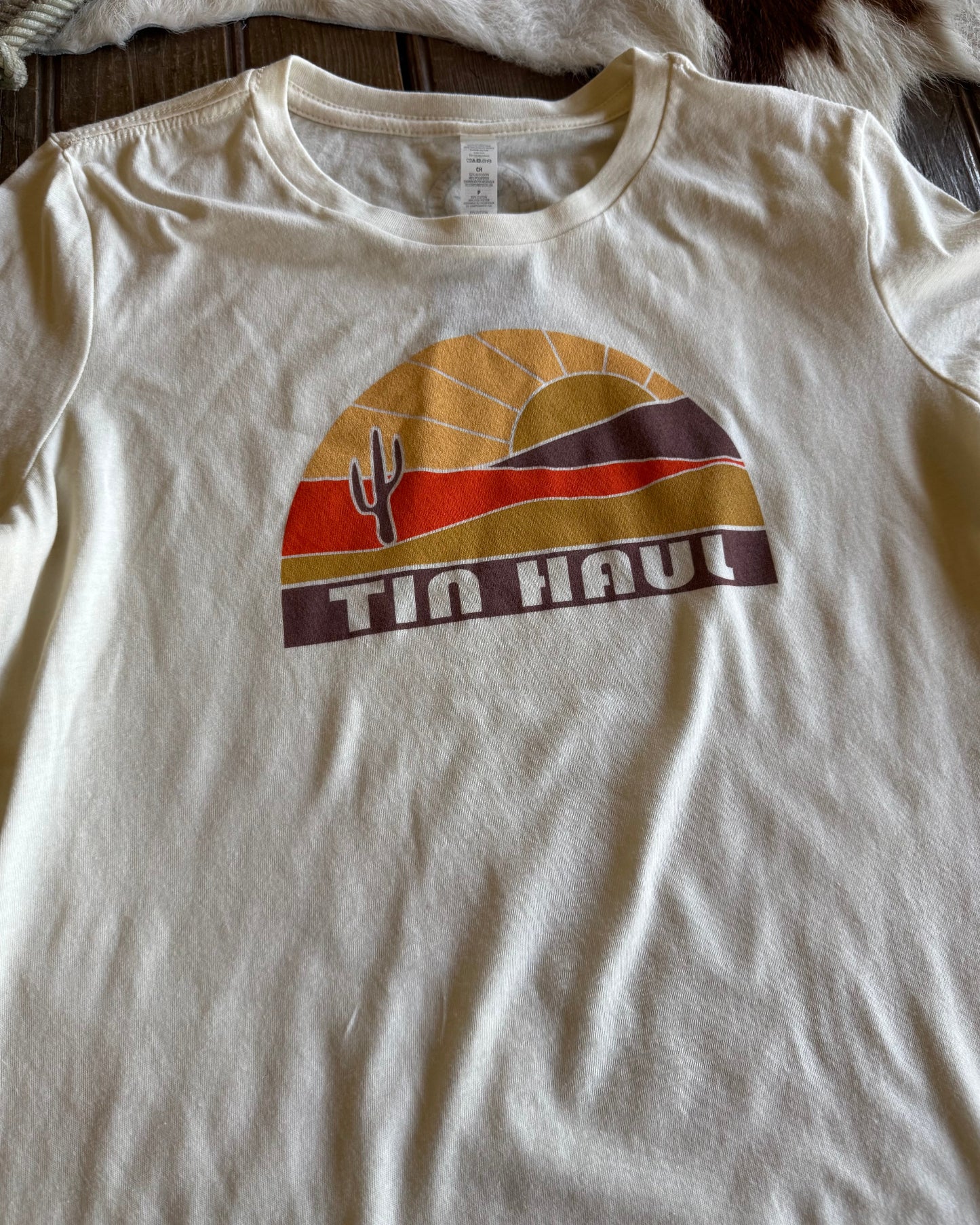 White t-shirt with a graphic design featuring a sunset and cactus, labeled 'Tin Haul', on a wooden surface.