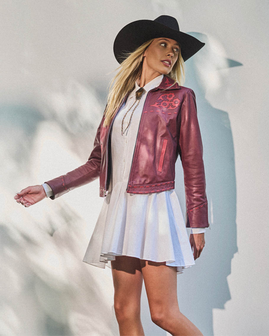Woman wearing a red leather jacket, white dress, and black hat against a light background