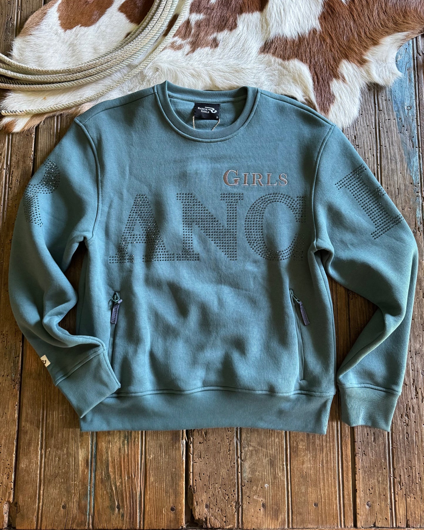 blue sweatshirt with 'RANCH GIRLS' text on a wooden surface with cowhide rug