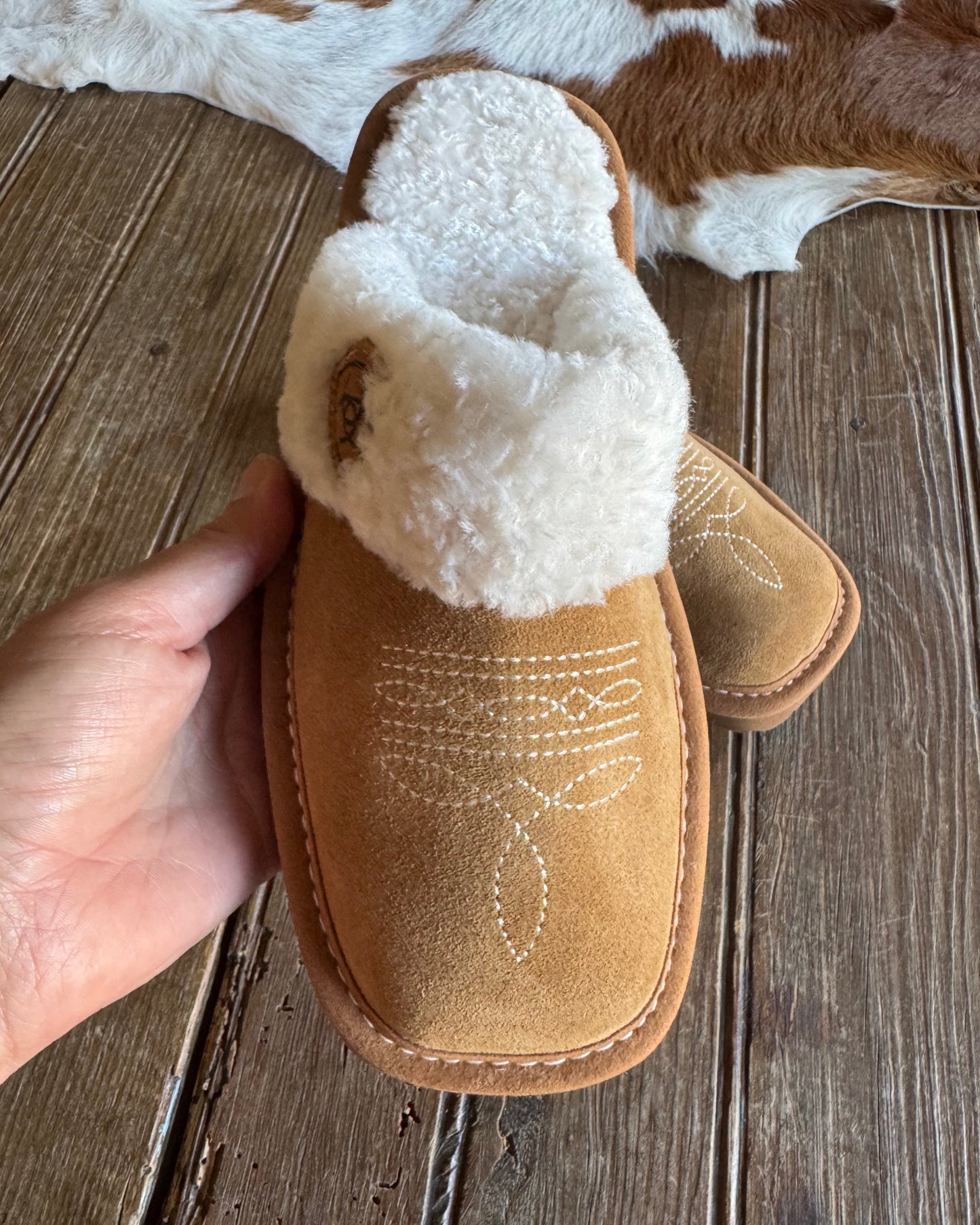 Brown slipper with white fur lining held by a hand on a wooden floor.