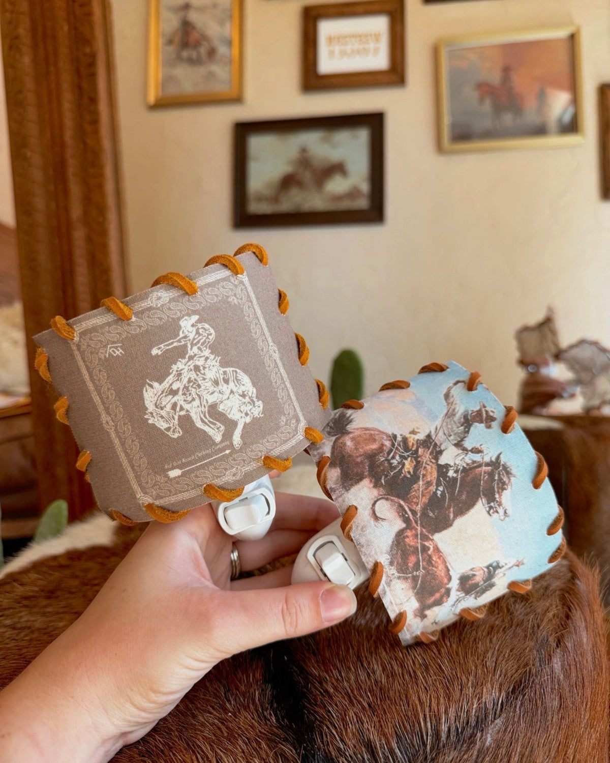 A hand holds two western-themed leather hair clips with white plastic spring clips, one featuring a white cowboy illustration and the other a western landscape print, against a rustic indoor background.