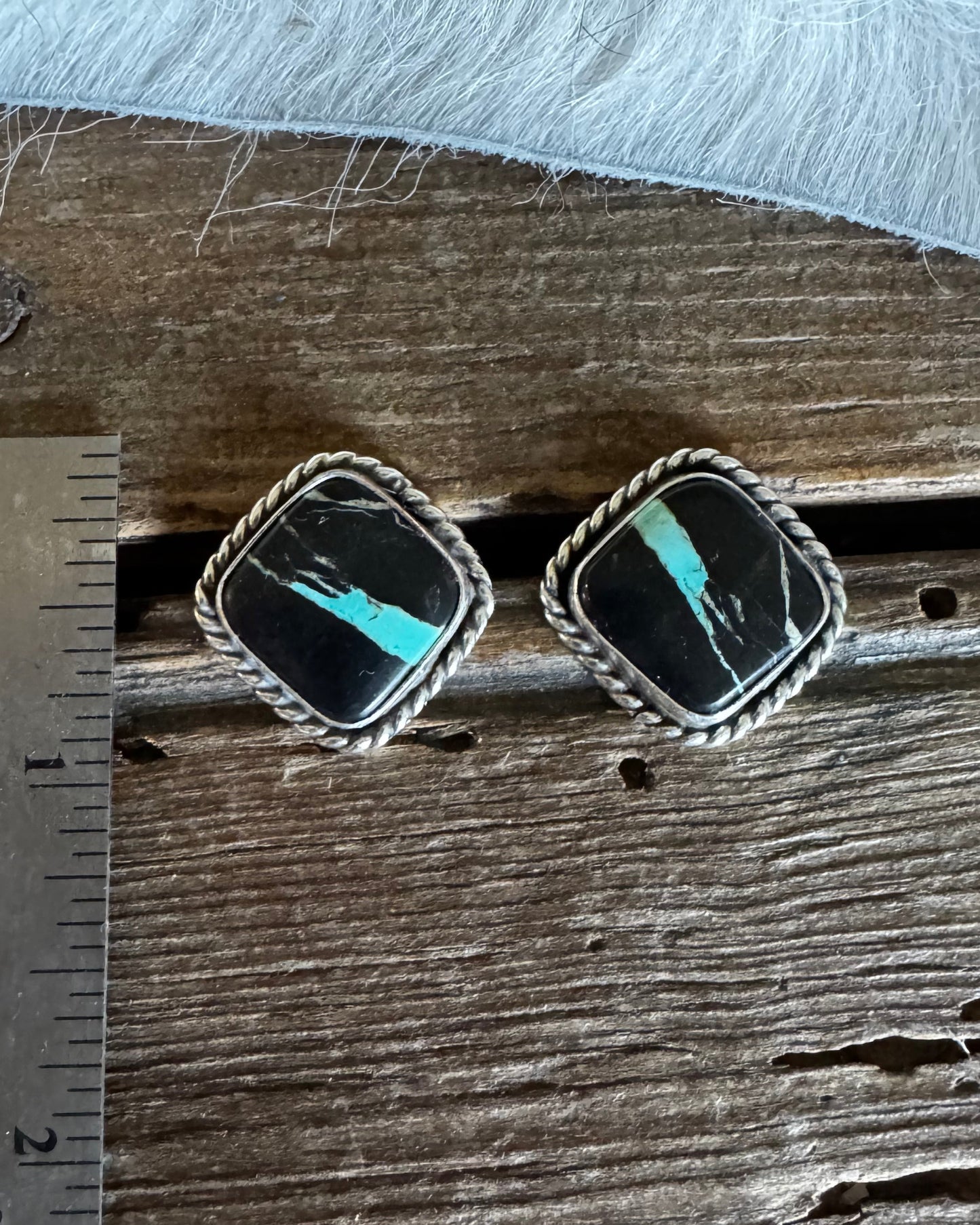 A pair of square silver-toned earrings with black inlay and turquoise-colored stone accents, displayed on a rustic wooden surface next to a metal ruler for scale.