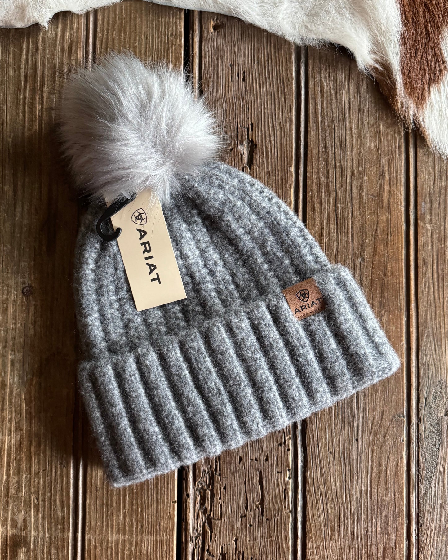 Gray knit beanie with pom-pom and Ariat tag on a wooden surface