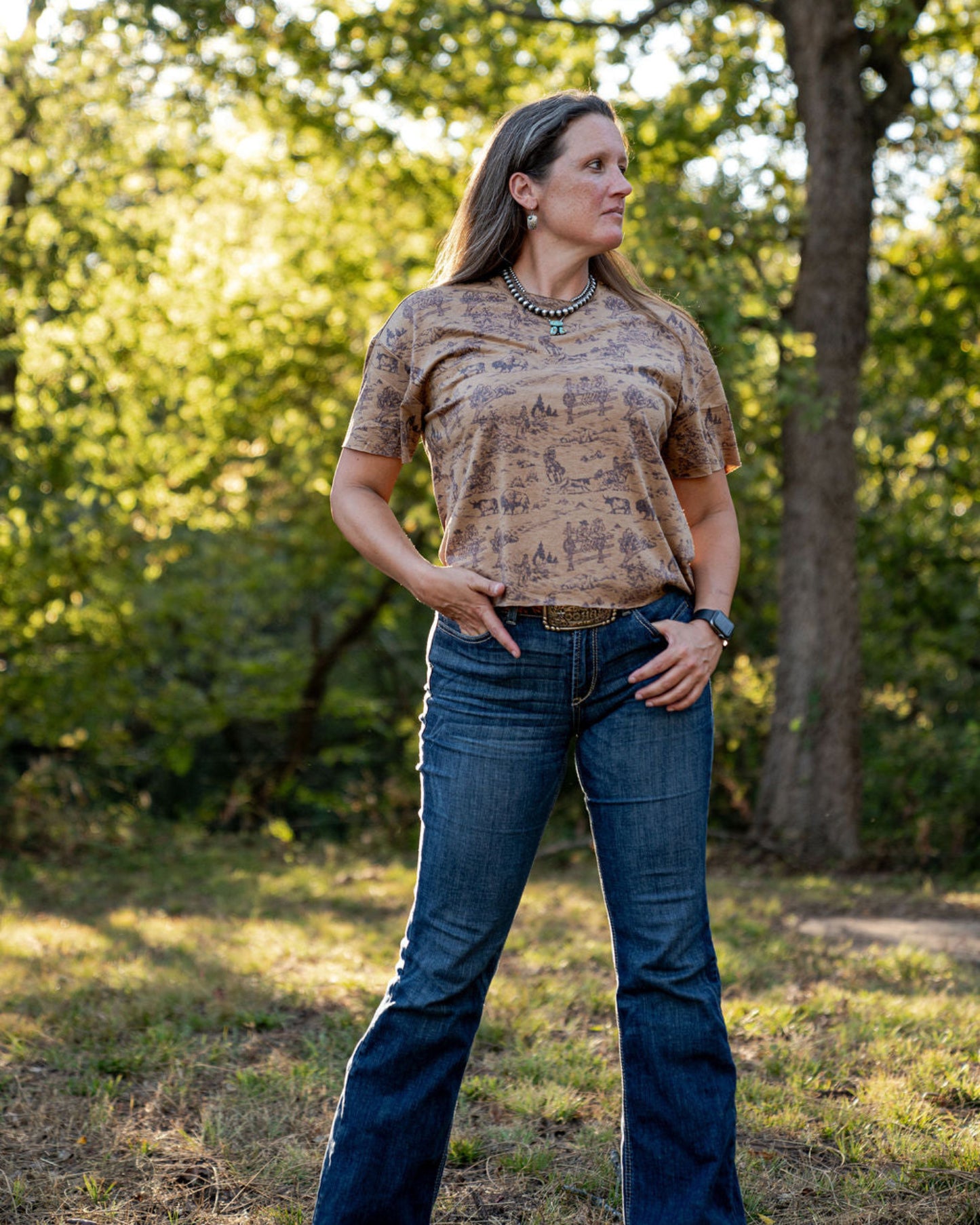 Woman wearing a patterned shirt and jeans standing in a natural setting with trees and grass.