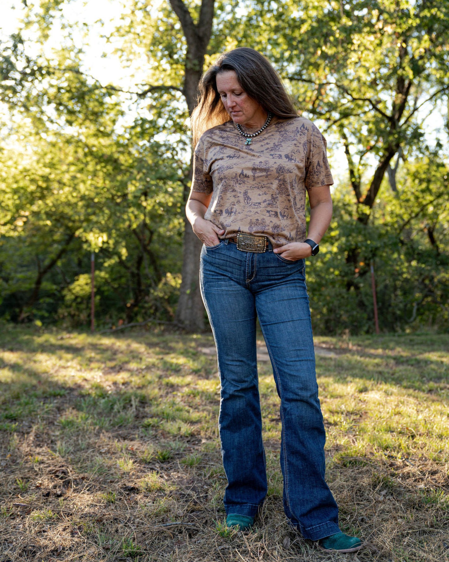Woman standing in a natural setting wearing a patterned top and blue jeans.