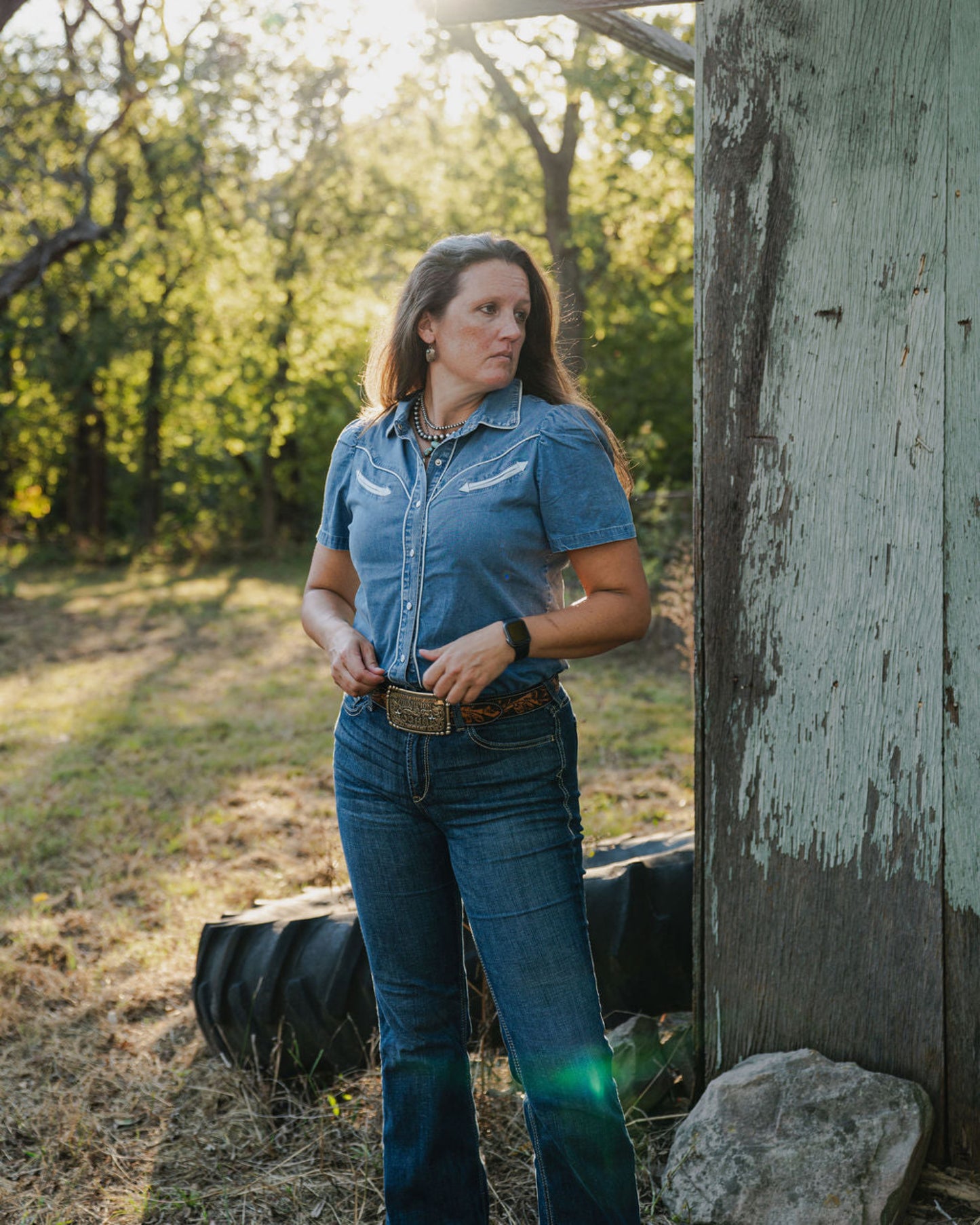 Woman in a blue denim shirt and jeans standing outdoors near a wooden structure with trees in the background.