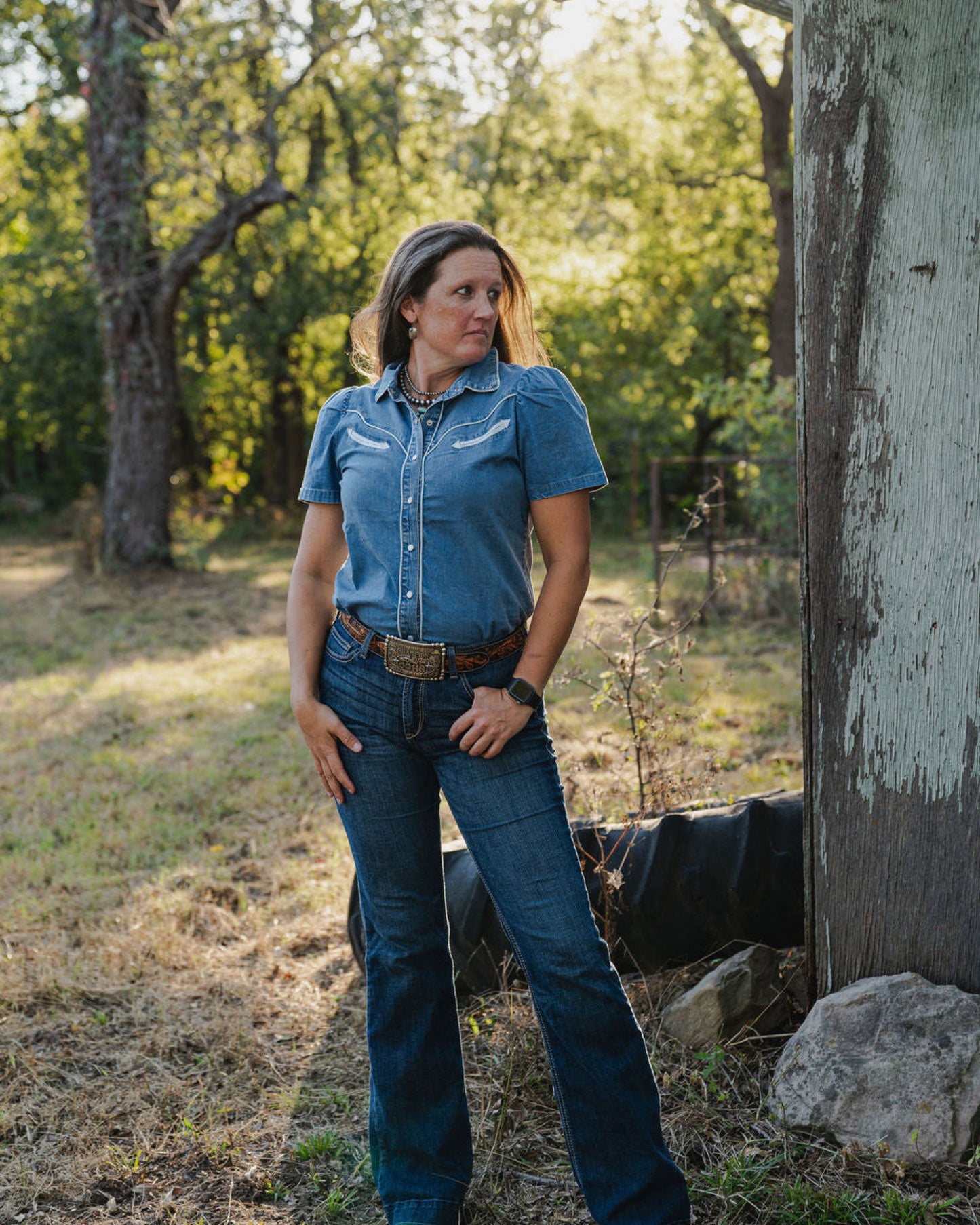 Woman in a blue shirt and jeans standing outdoors with trees and sunlight in the background