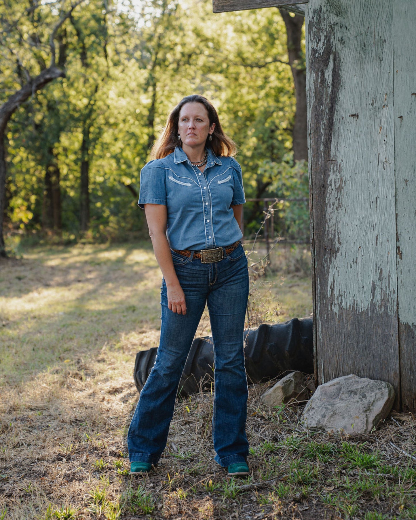 Woman in a denim outfit standing in a rural setting with trees and a wooden structure.