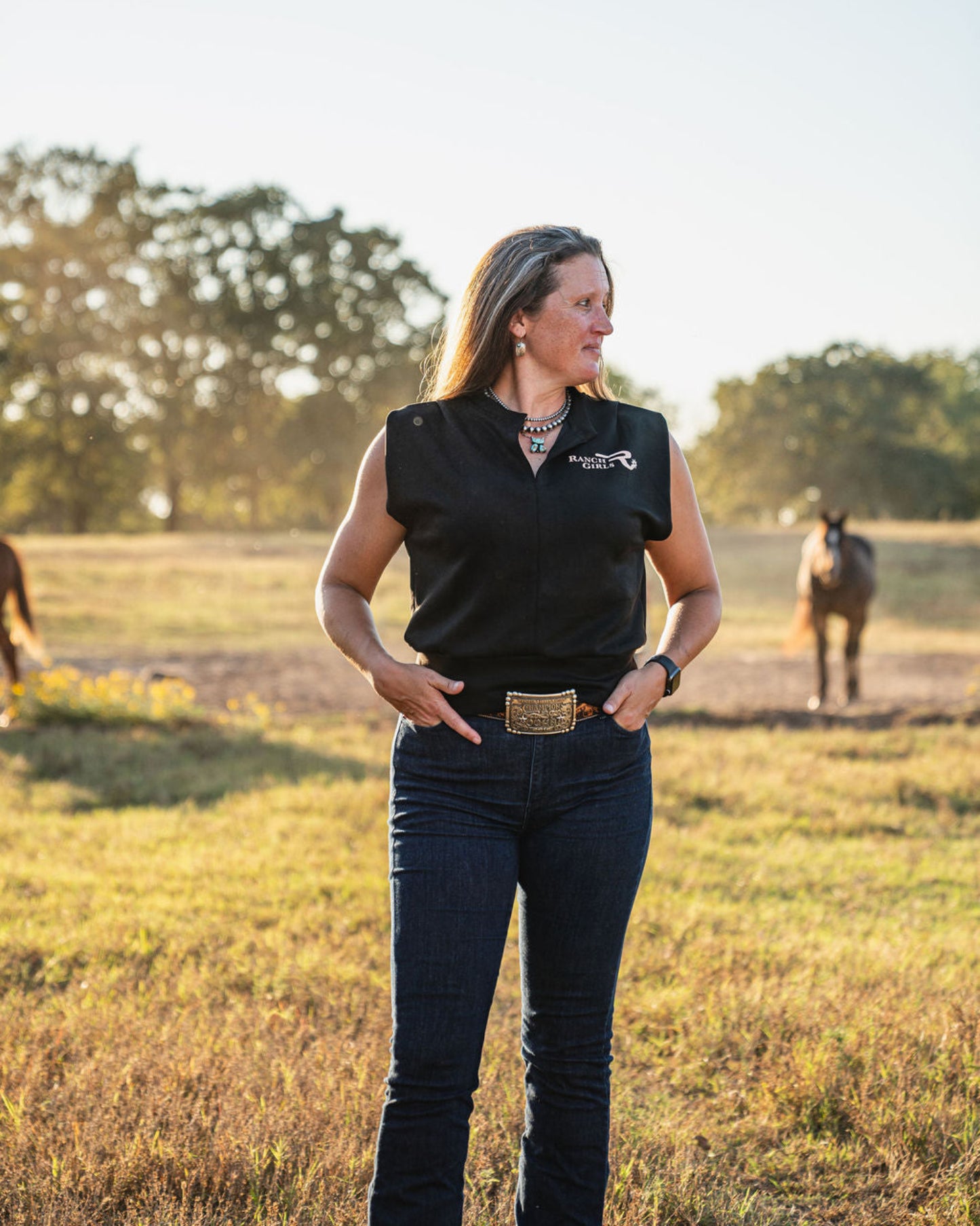 Woman standing in a field with horses, wearing a black shirt with a logo.