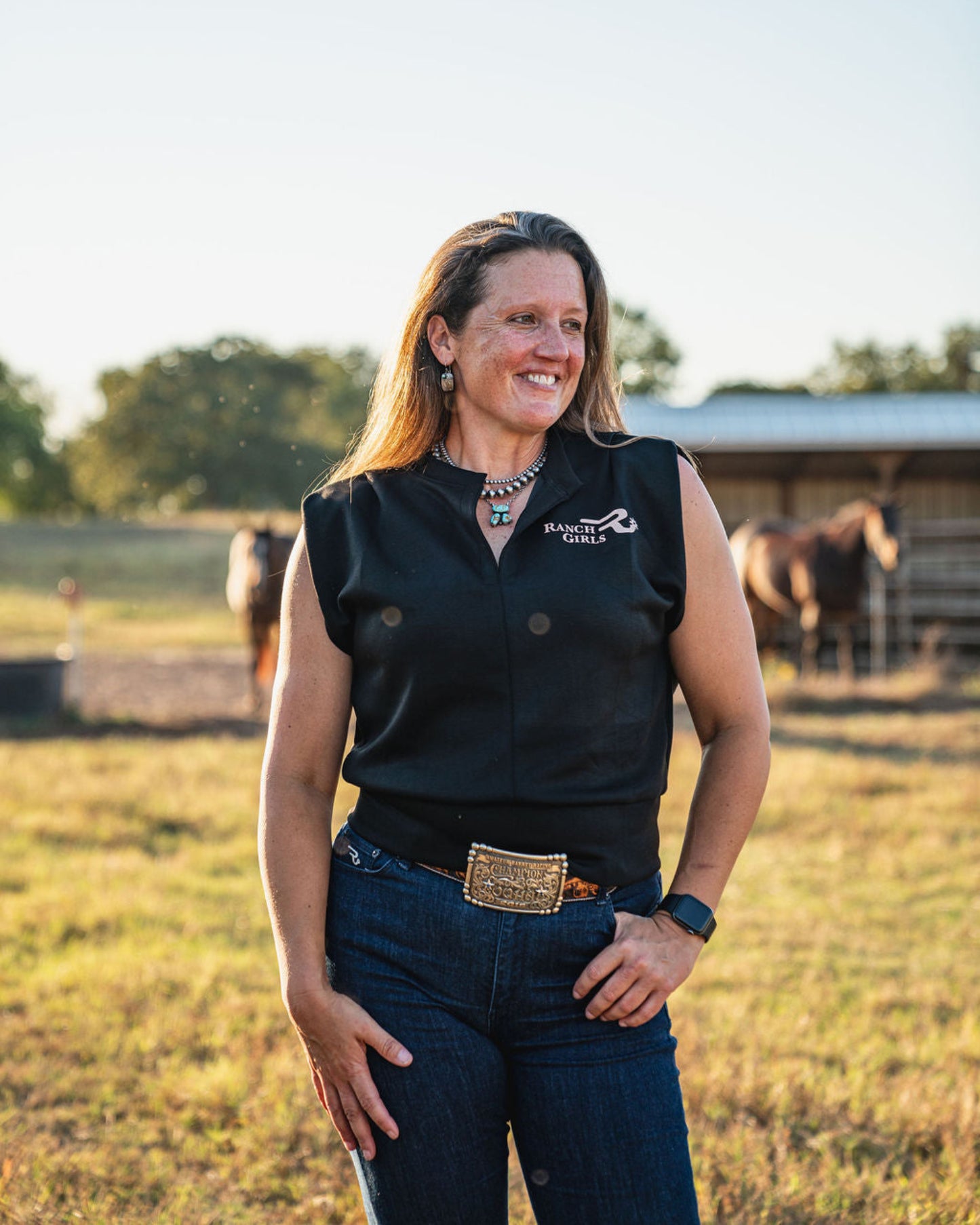 Woman wearing a black sleeveless shirt with a logo, standing in an outdoor setting with horses in the background.