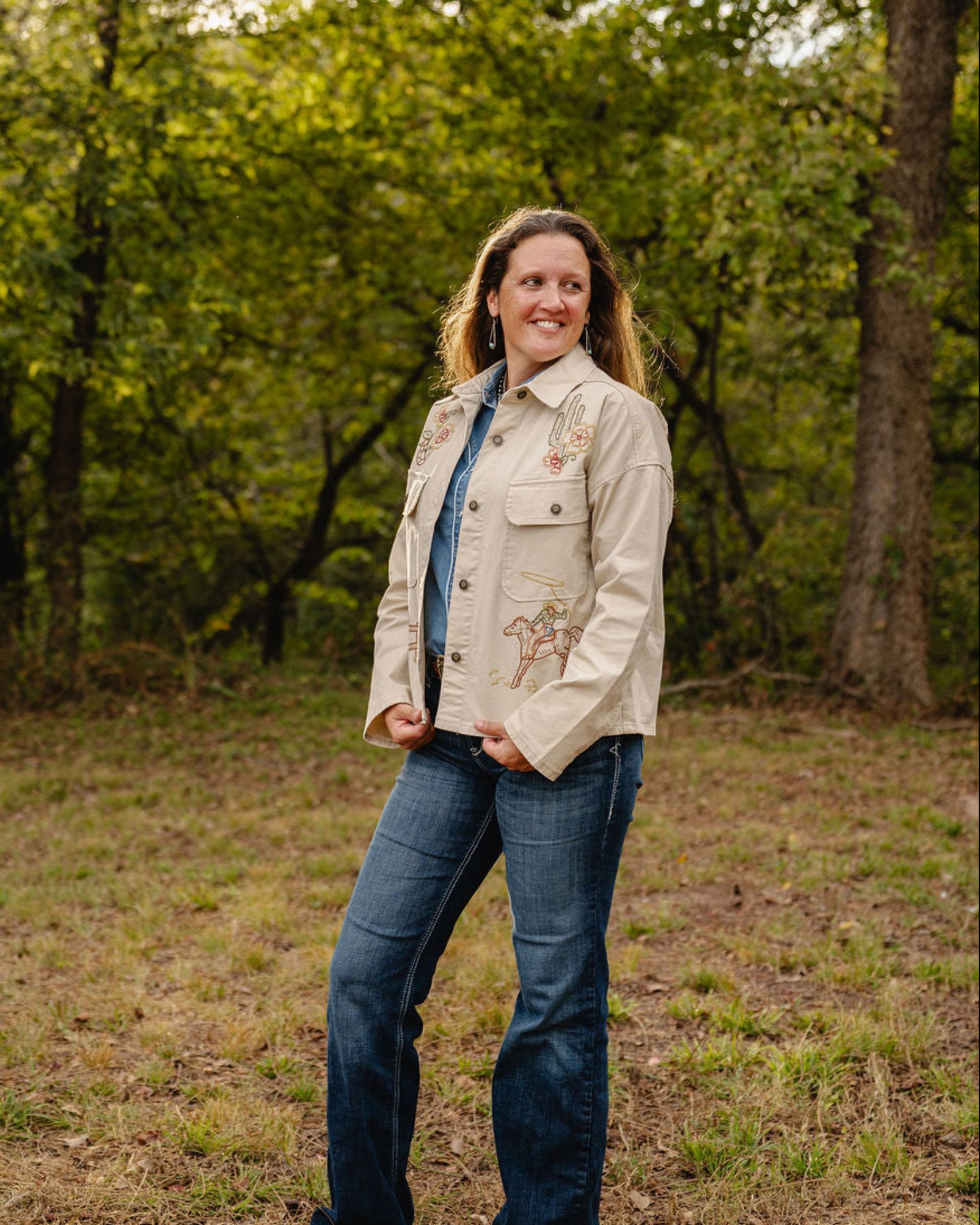 Woman standing in a forested area wearing a beige jacket with a floral design.
