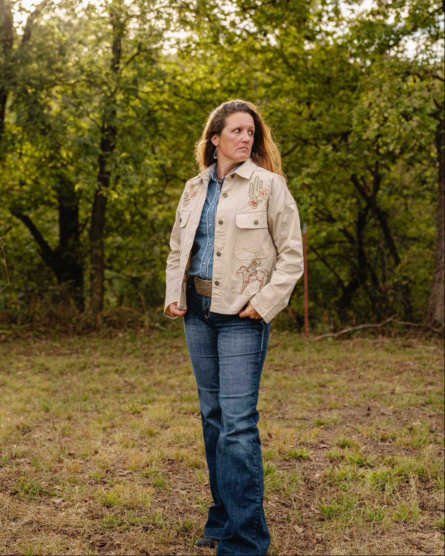 Woman standing in a forested area wearing a beige jacket and blue jeans.