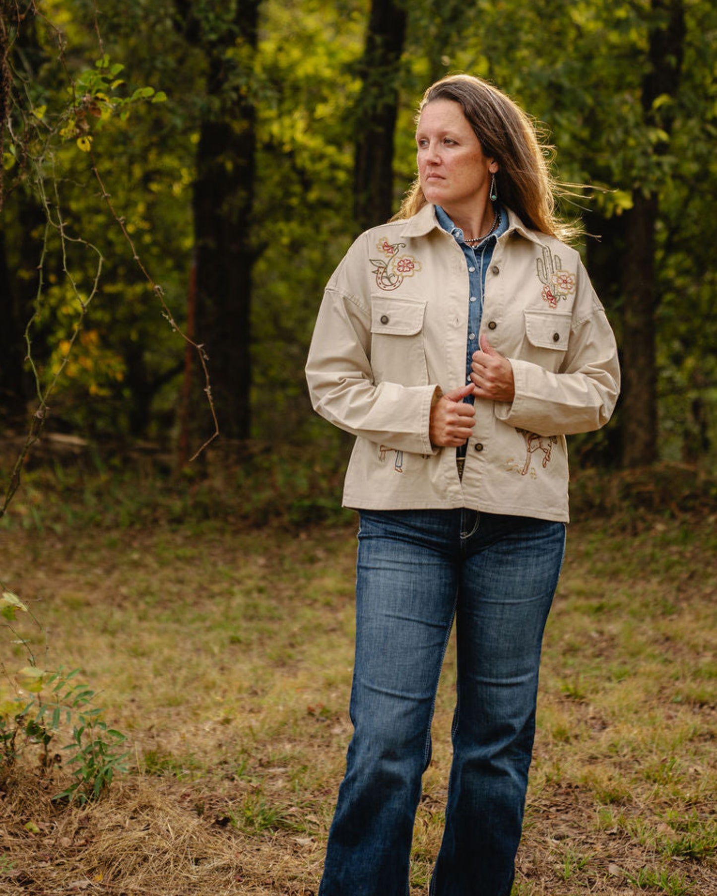 Woman standing in a forested area wearing a beige jacket and blue jeans.